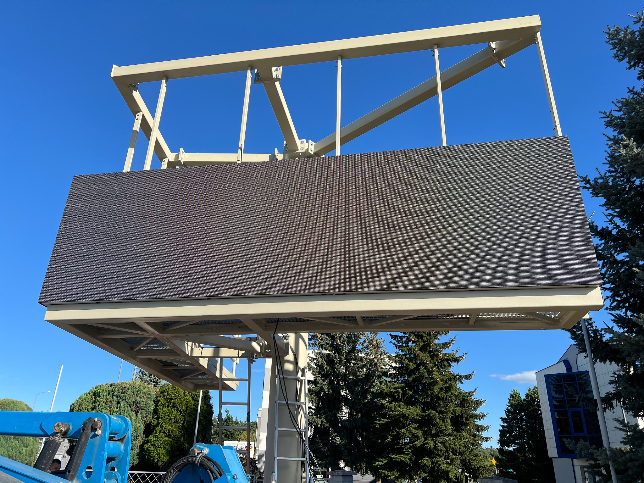 Zewnętrzny ekran LED na konstrukcji stalowej, widok z dołu na tle błękitnego nieba i zieleni drzew. Widoczny fragment podnośnika koszowego.