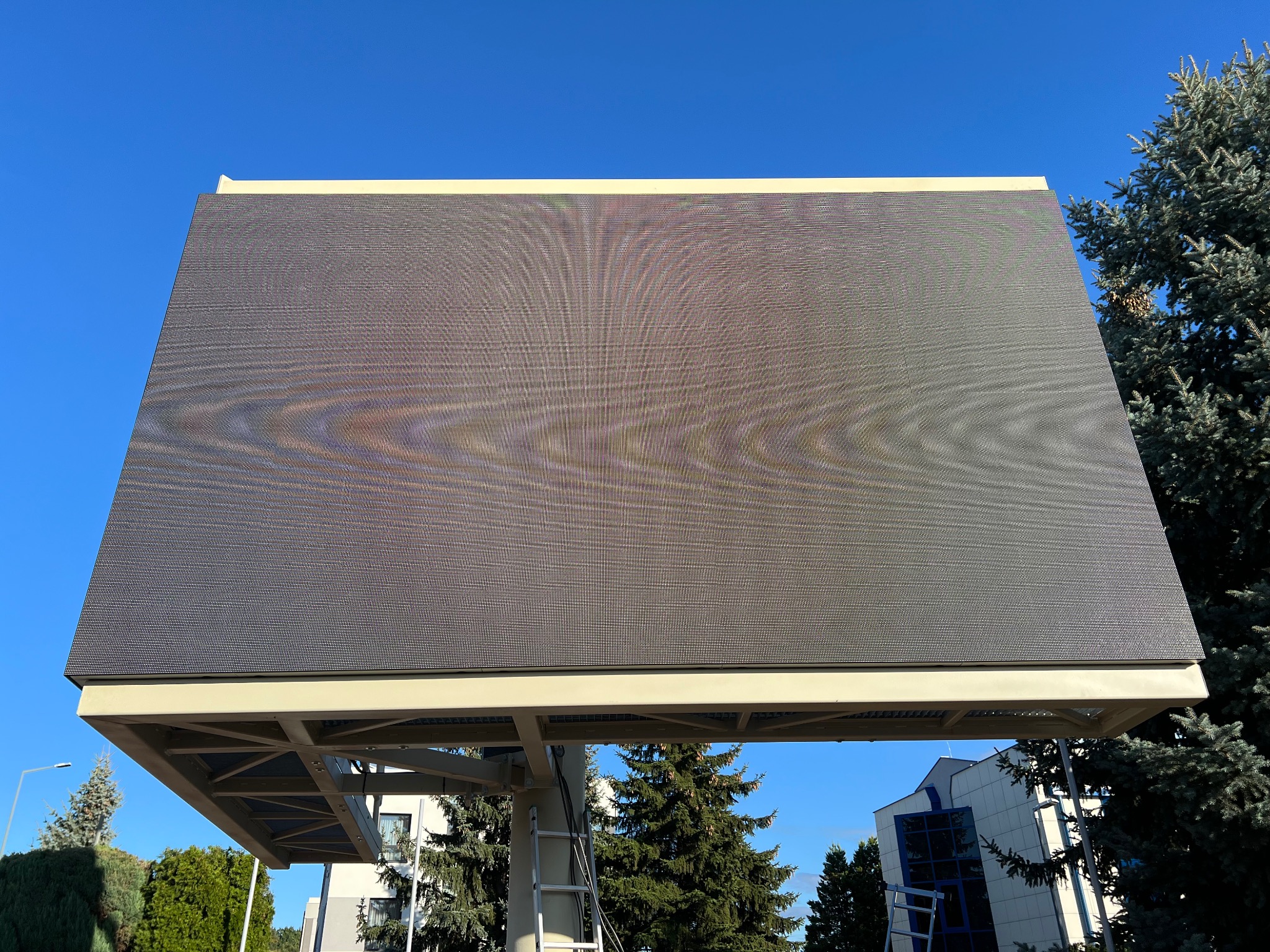 Duży ekran LED na konstrukcji stalowej, widziany z dołu na tle błękitnego nieba i zielonych drzew. Widoczna drabina sugeruje prace montażowe lub serwisowe.