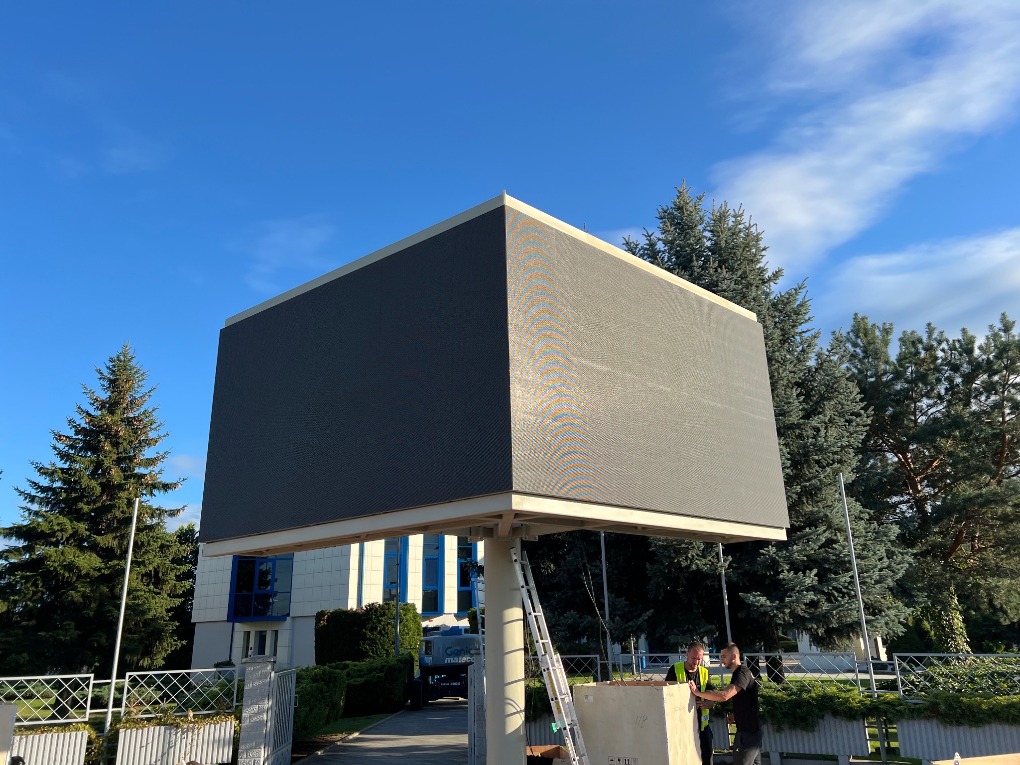 Wysoki ekran LED na konstrukcji, montowany przez dwóch mężczyzn w kamizelkach odblaskowych. W tle budynek z niebieskimi oknami i drzewa, błękitne niebo.