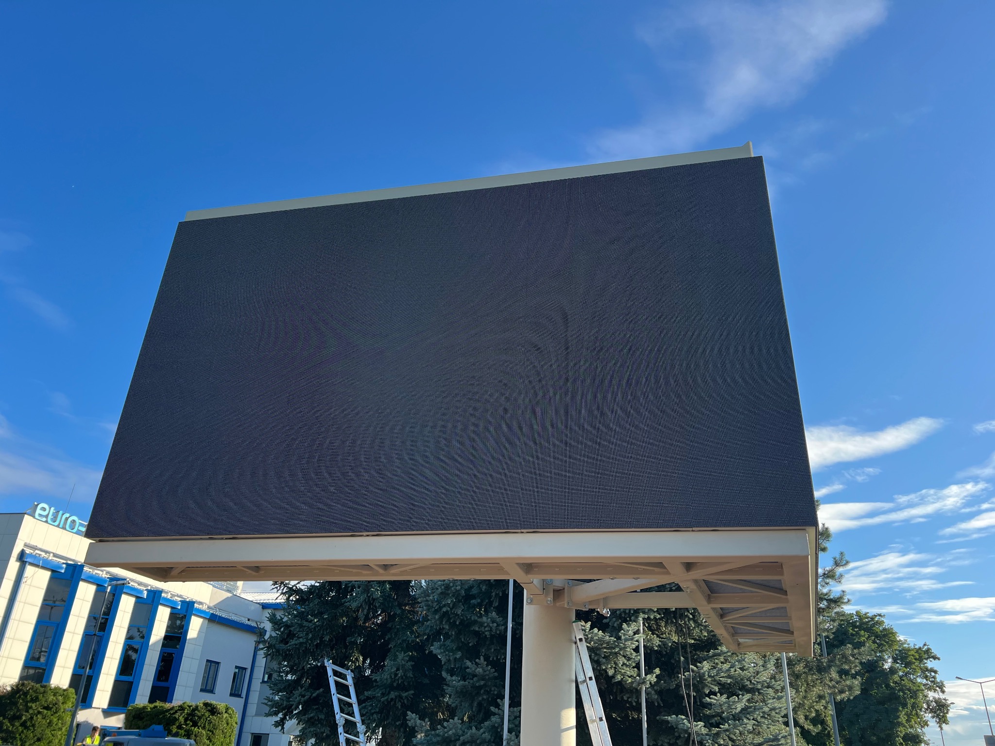 Duży ekran LED na konstrukcji stalowej, widoczny z dołu na tle błękitnego nieba. Dwie drabiny oparte o konstrukcję. Budynek w tle.