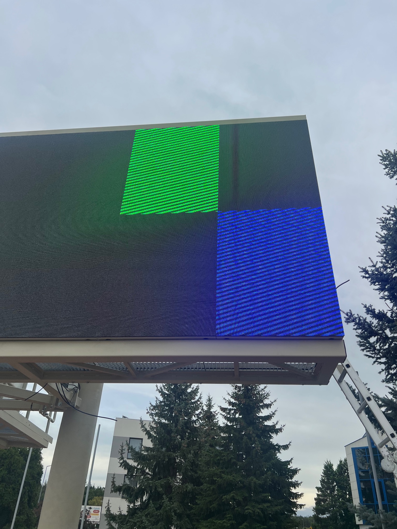 Włączony, duży ekran LED z zielonym i niebieskim kwadratem, widziany z dołu. Drabina oparta o konstrukcję. Tło stanowią drzewa i budynek.