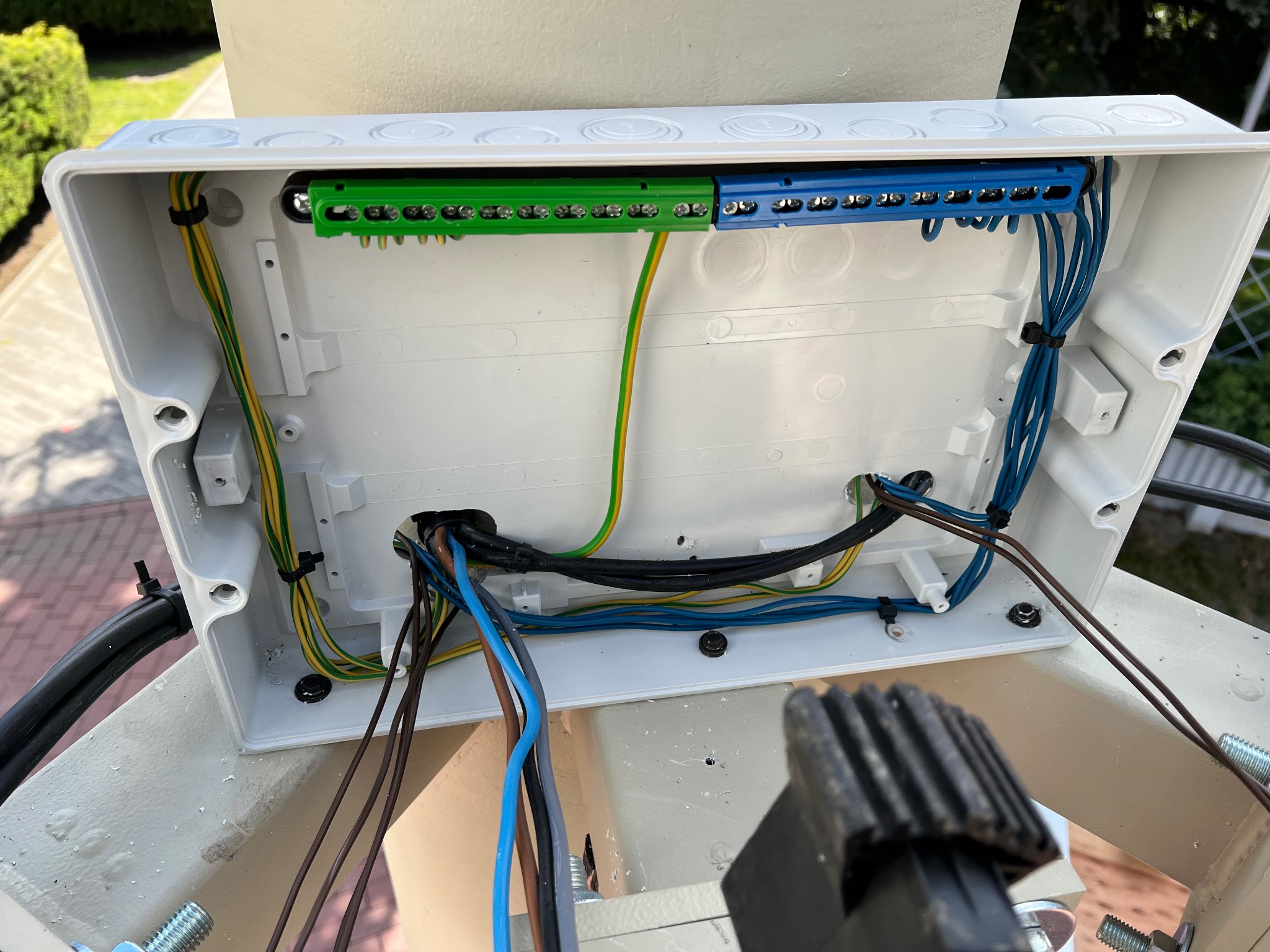 Otwarta, biała puszka elektryczna z podłączonymi przewodami w kolorach: niebieskim, brązowym, czarnym i żółto-zielonym. Widoczne listwy zaciskowe w kolorze zielonym i niebieskim.