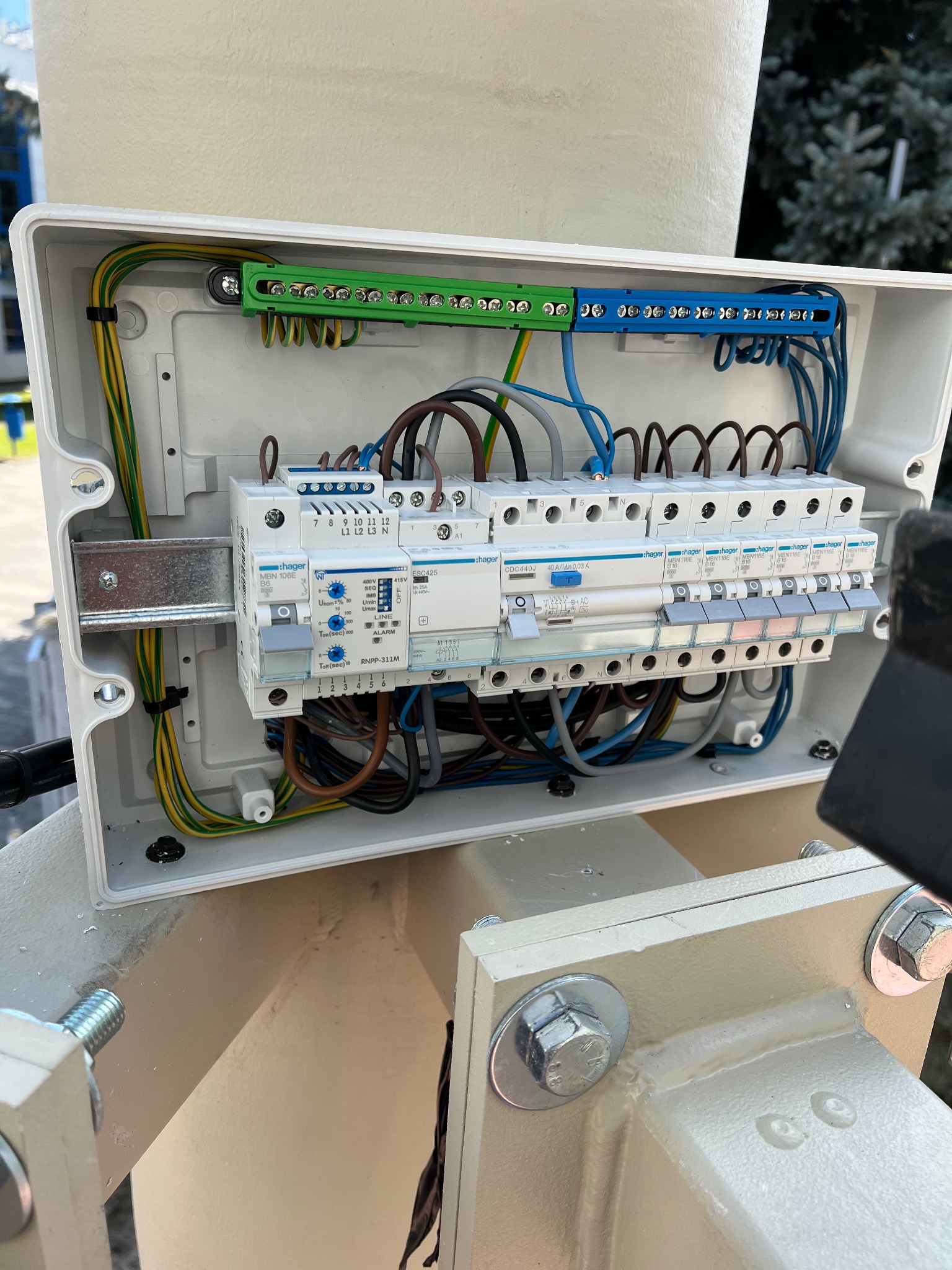 Otwarta skrzynka elektryczna z bezpiecznikami Hager, widoczne przewody w kolorach: żółto-zielonym, brązowym, niebieskim i szarym. Element instalacji zamontowany na konstrukcji.