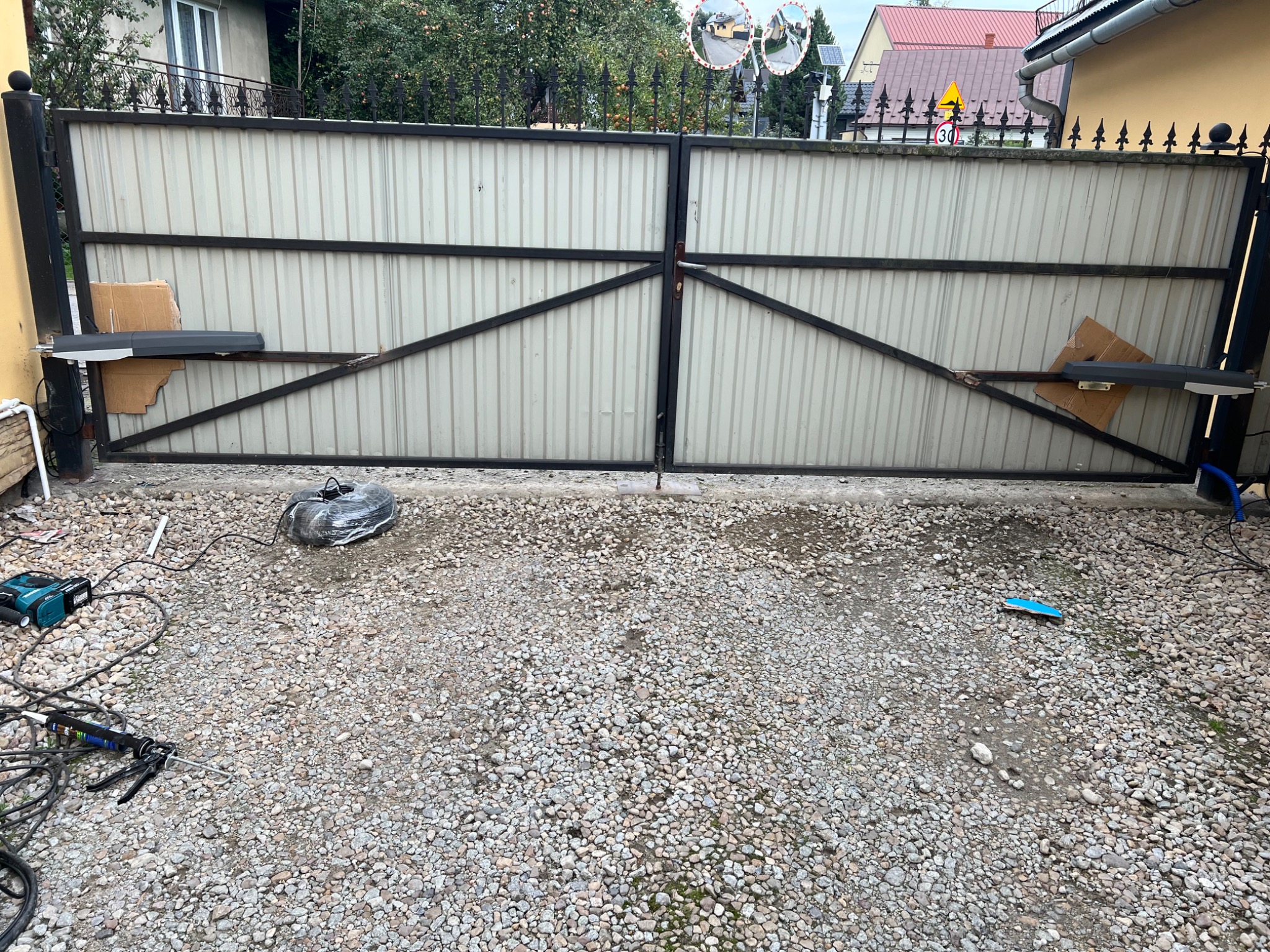 Biała, metalowa brama dwuskrzydłowa z czarną konstrukcją w trakcie montażu automatyki. Na ziemi narzędzia i kabel. Widoczne domy w tle.