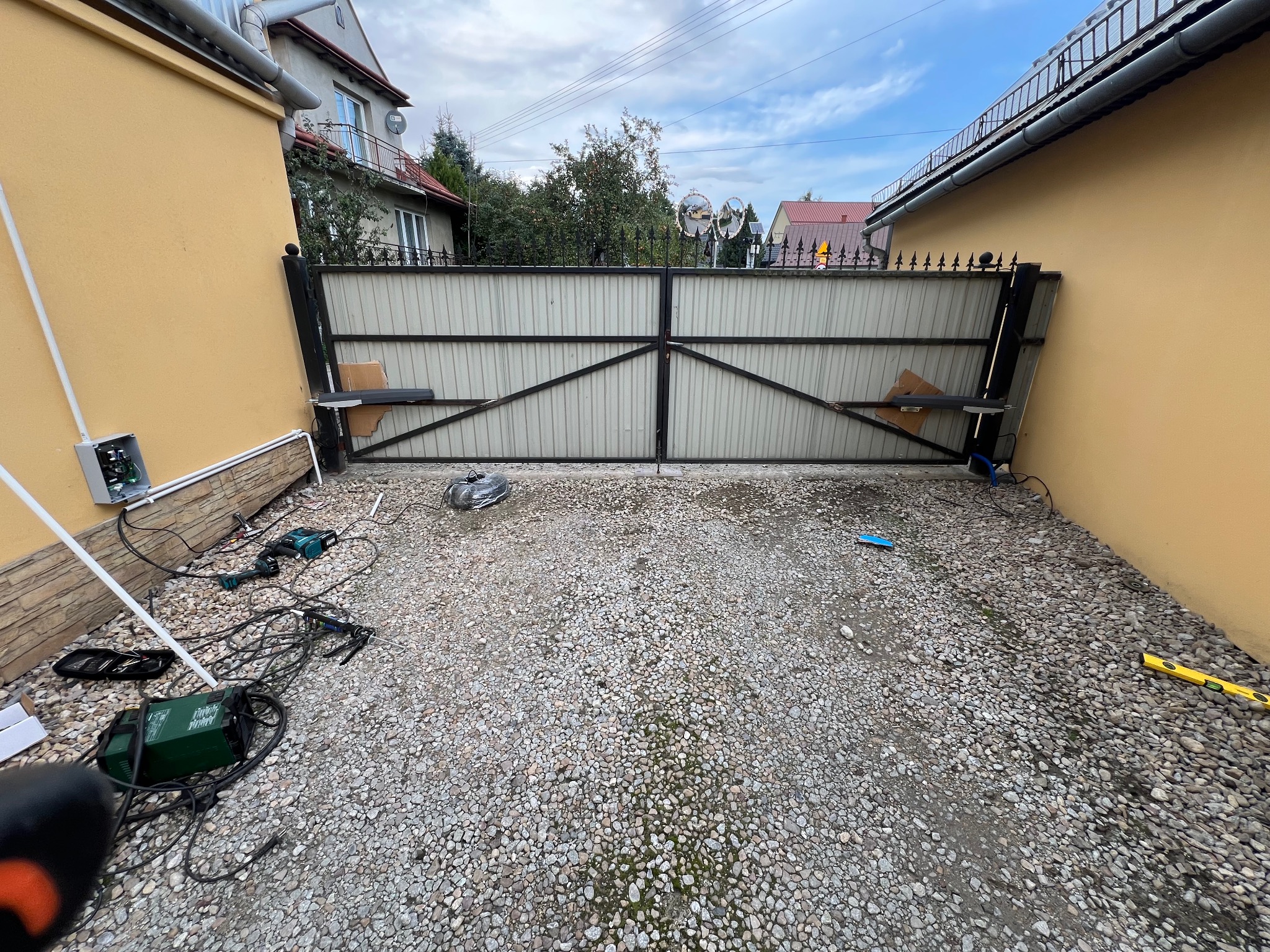 Montaż automatycznej bramy wjazdowej na posesję. Widoczne narzędzia i elementy instalacyjne na żwirowym podłożu. Brama częściowo zamontowana, z widocznym mechanizmem.