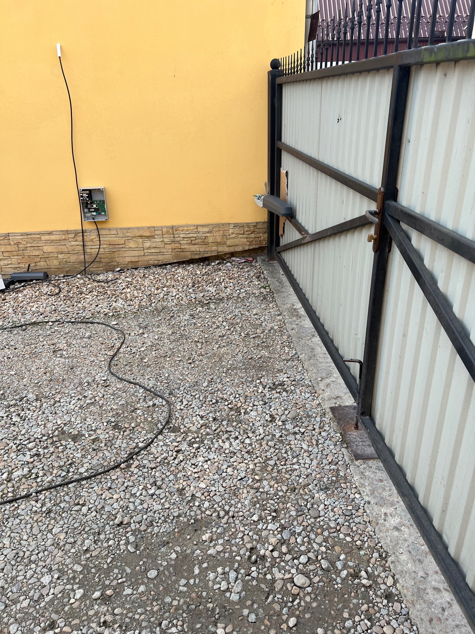 Brama przesuwna z automatyką, widoczny mechanizm napędu i skrzynka sterująca na żółtej ścianie. Kabel zasilający na żwirowym podłożu.