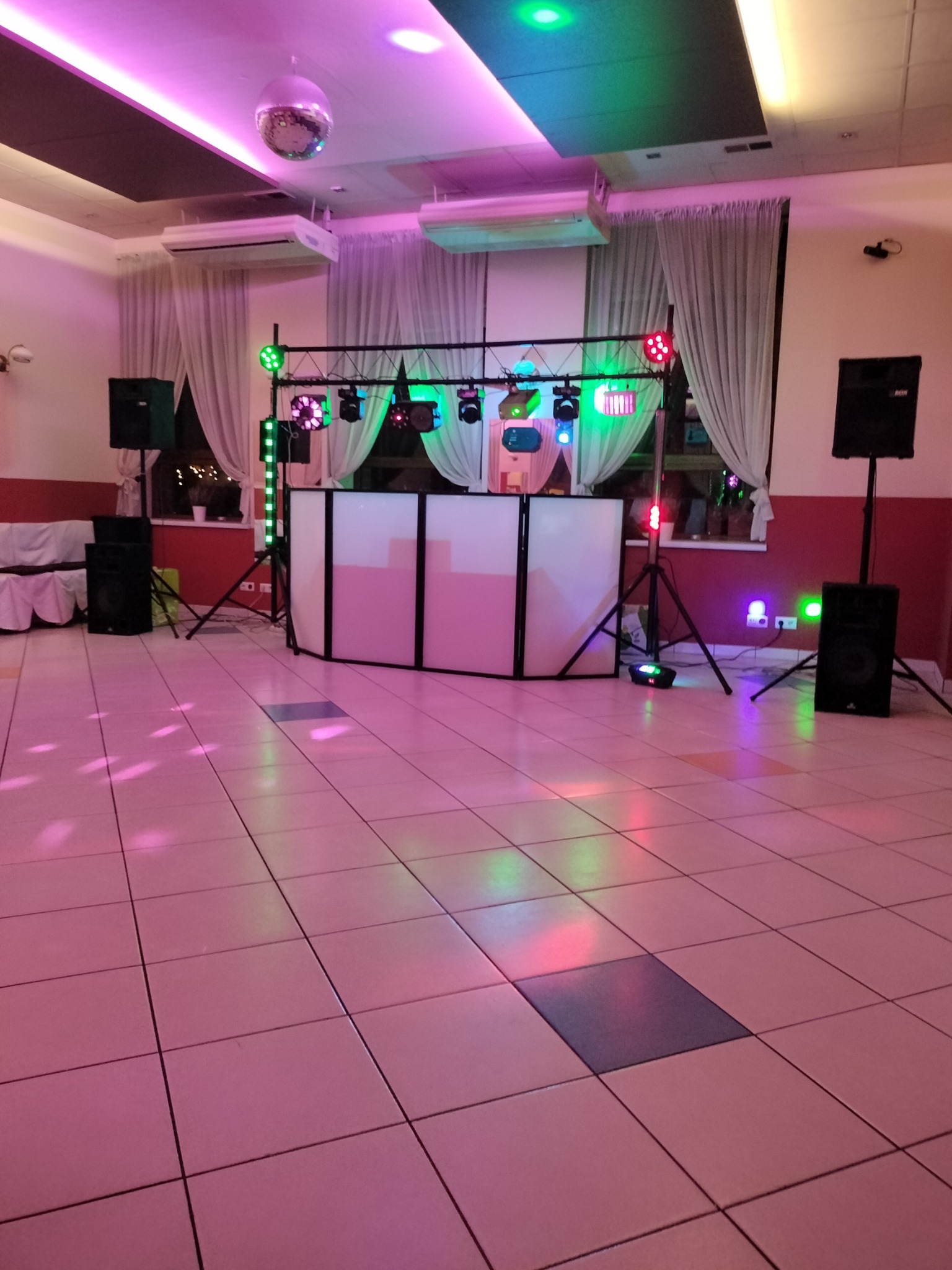 Rozstawiony sprzęt nagłośnieniowy i oświetleniowy w sali z parkietem, widoczny biały panel DJ-ski, kolumny głośnikowe, reflektory i kula dyskotekowa pod sufitem.