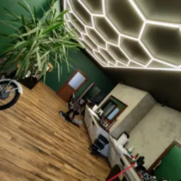 Wnętrze salonu fryzjerskiego z nowoczesnym oświetleniem sufitowym w kształcie plastra miodu, drewnianą podłogą, zielonymi ścianami, fotelem fryzjerskim i rośliną doniczkową przy motocyklu.