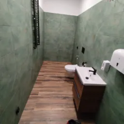 Wąska łazienka po remoncie z zielonymi płytkami ściennymi, drewnopodobną podłogą, wiszącą szafką z umywalką, czarną armaturą i grzejnikiem drabinkowym. Na podłodze leżą robocze buty.