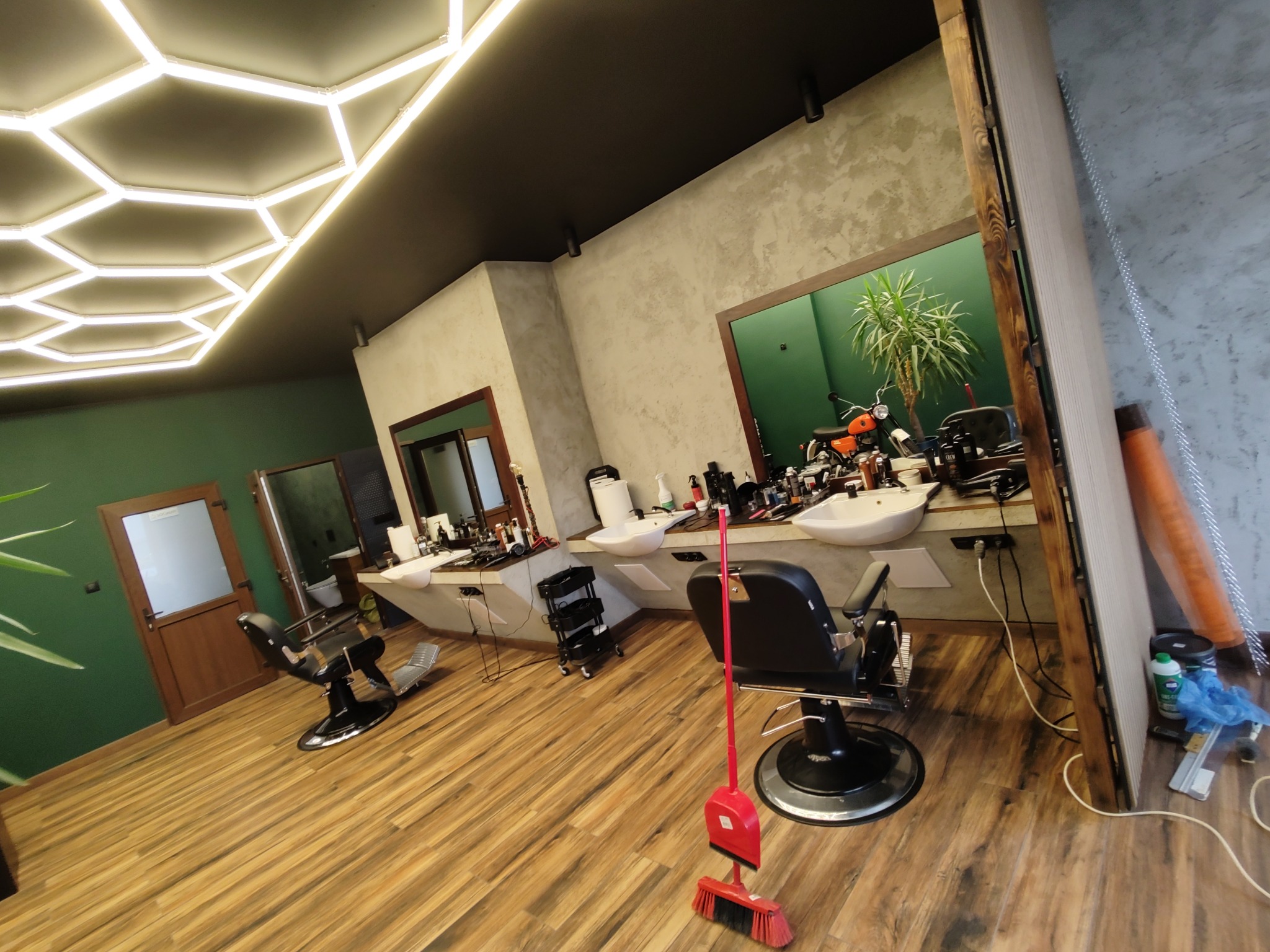 Wnętrze salonu fryzjerskiego z dwoma stanowiskami, fotelami barberskimi, umywalkami, narzędziami fryzjerskimi, nietypowym oświetleniem sufitowym w kształcie plastra miodu oraz czerwoną zmiotką...