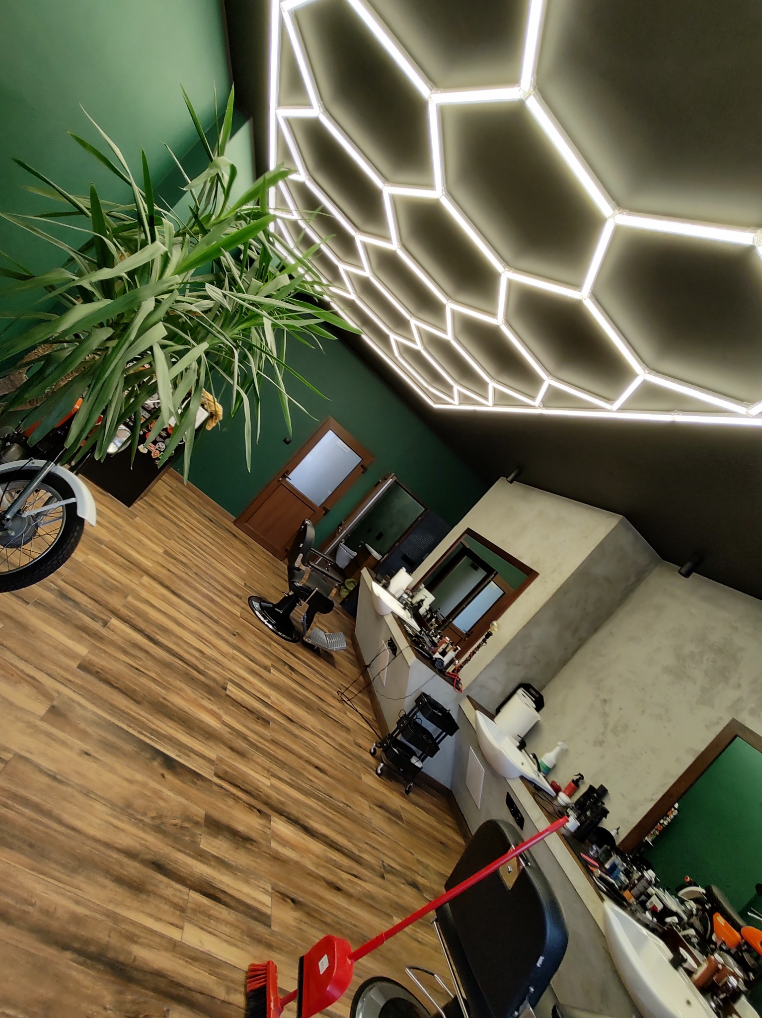 Wnętrze salonu fryzjerskiego z nowoczesnym oświetleniem sufitowym w kształcie plastra miodu, drewnianą podłogą, zielonymi ścianami, fotelem fryzjerskim i rośliną doniczkową przy motocyklu.