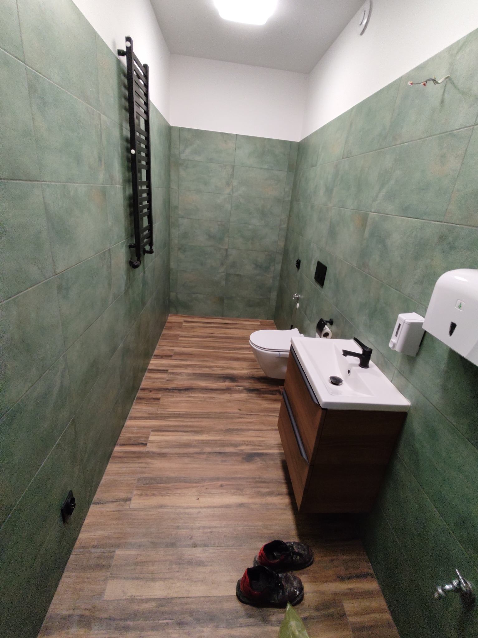 Wąska łazienka po remoncie z zielonymi płytkami ściennymi, drewnopodobną podłogą, wiszącą szafką z umywalką, czarną armaturą i grzejnikiem drabinkowym. Na podłodze leżą robocze buty.