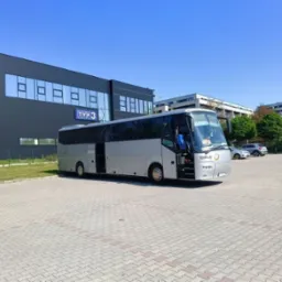 Srebrny autokar turystyczny z otwartymi drzwiami na parkingu przed budynkiem TVP3 w słoneczny dzień.