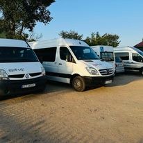 Flota białych busów transportowych różnych marek zaparkowana na piaszczystym placu w słoneczny dzień. Widoczny fragment zabudowań w tle.