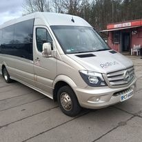 Złoty bus Mercedes Sprinter z przyciemnianymi szybami, zaparkowany na betonowym placu przed budynkiem z czerwonym dachem.