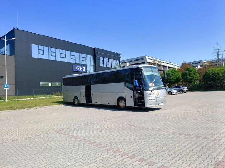 Srebrny autokar turystyczny z otwartymi drzwiami na parkingu przed budynkiem TVP3 w słoneczny dzień.