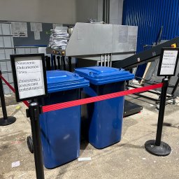 WIELOBRANŻOWA FIRMA HANDLOWA ANETA KASPRZAK WALDEMAR KASPRZAK ROBERT KASPRZAK SPÓŁKA JAWNA - Dwa niebieskie pojemniki na dokumenty przed przemysłową niszczarką, oddzielone czerwoną taśmą. Na podłodze widoczne resztki papieru, a w tle sterta dokumentów gotowych do zniszczenia.