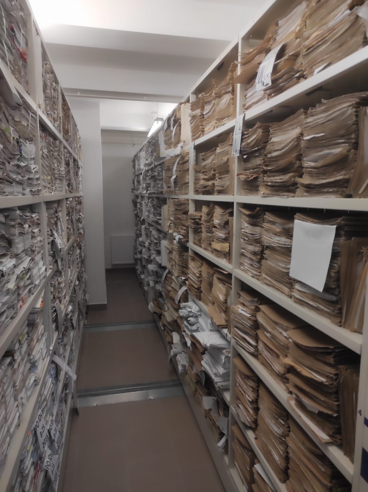 Archiwalne regały wypełnione dokumentami w teczkach, widok z wąskiego korytarza, oświetlenie jarzeniowe, brak osób.