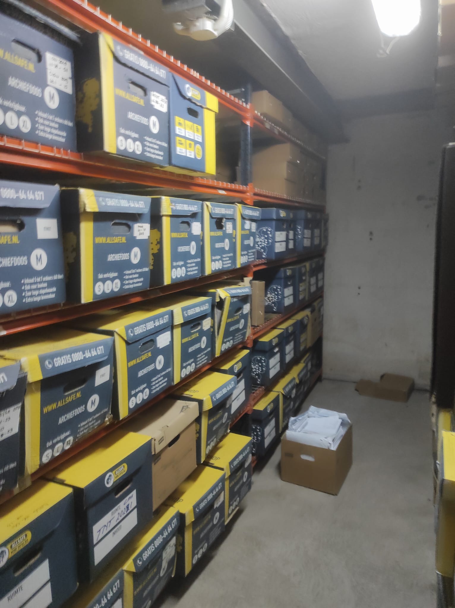 Magazyn z rzędami półek wypełnionych kartonami do archiwizacji dokumentów, niektóre z nich są wypełnione po brzegi dokumentami.