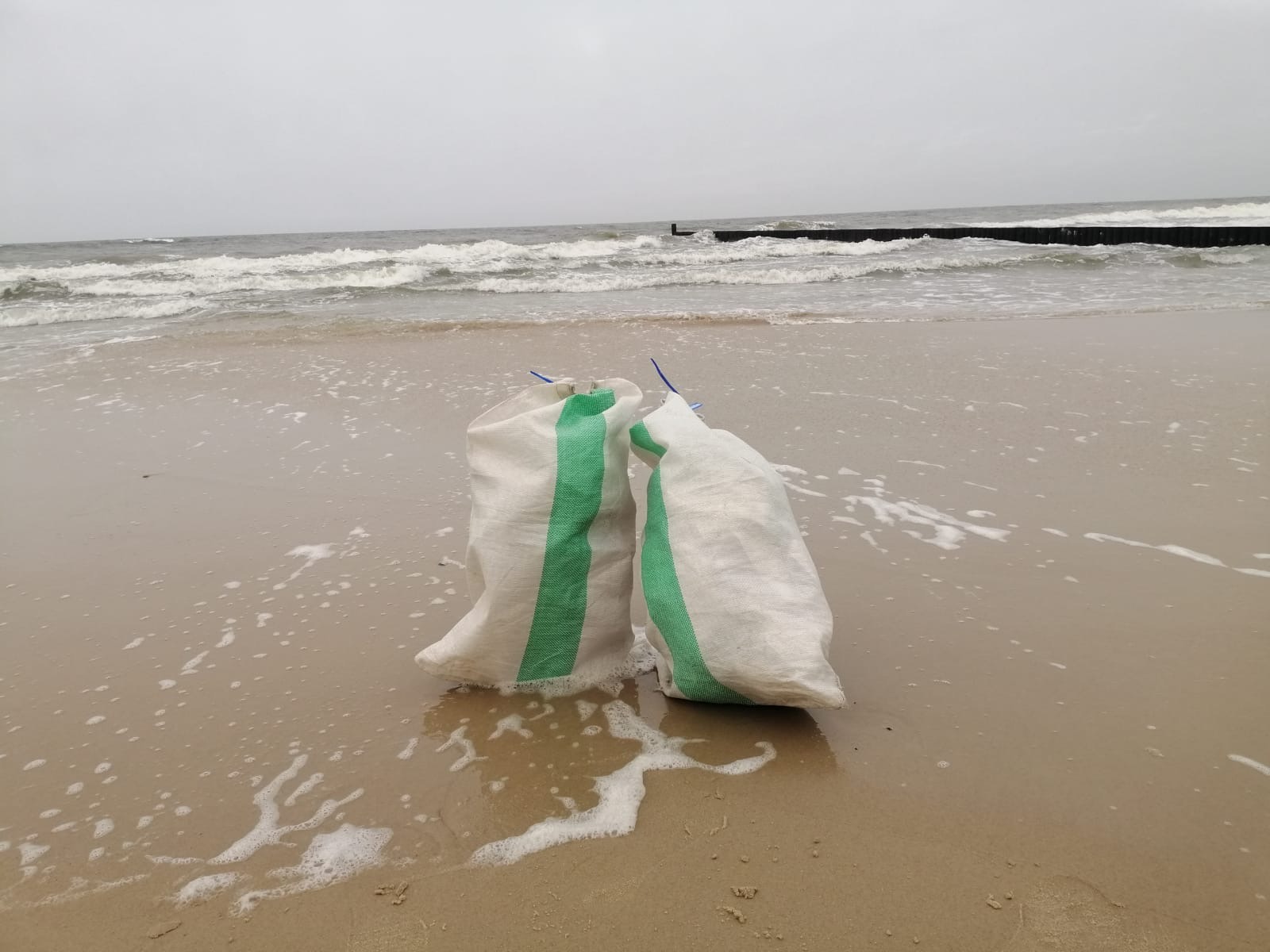 Dwa worki z zielonymi pasami stoją na mokrym piasku plaży, w tle morze i falochron.