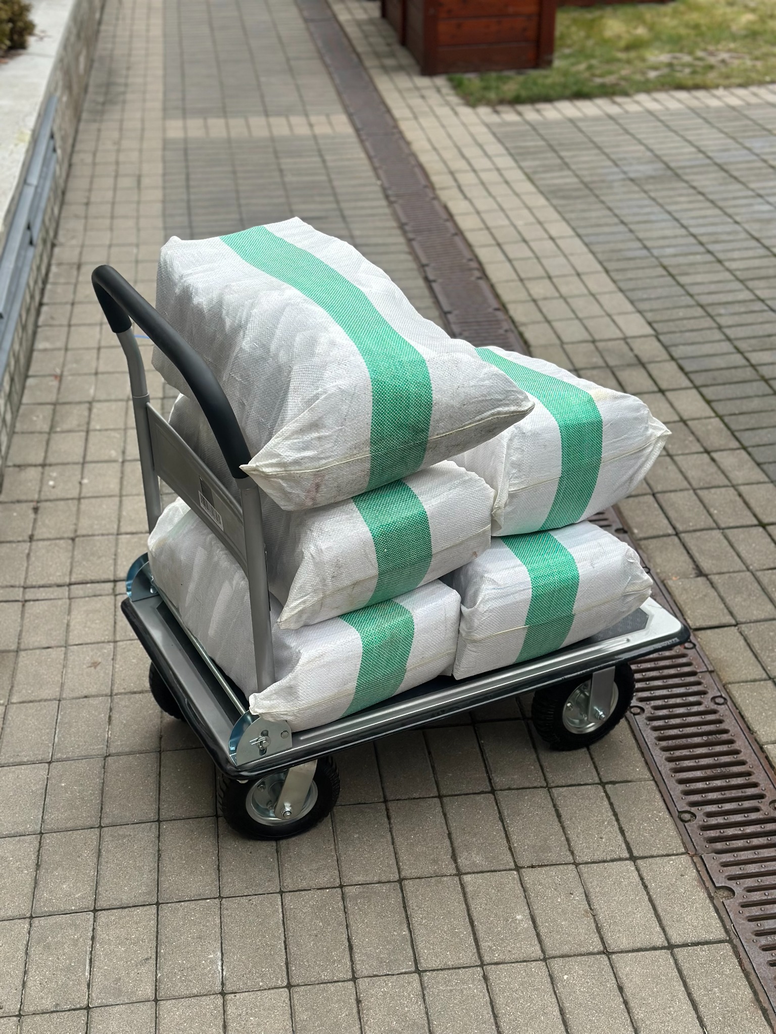 Wózek transportowy z workami z zawartością zabezpieczoną zielonymi pasami, stojący na kostce brukowej obok kratki ściekowej.