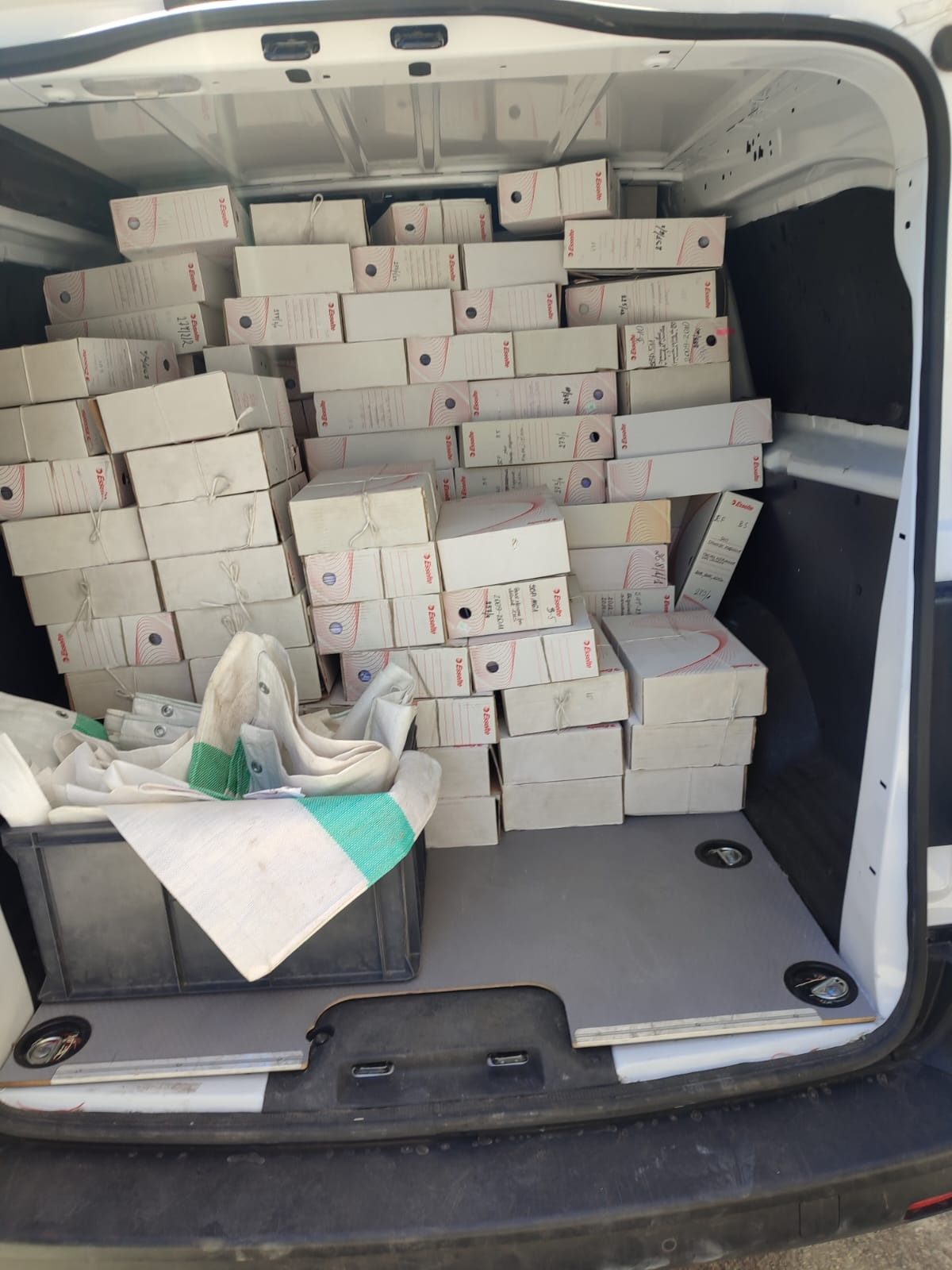 Wnętrze furgonetki wypełnionej po brzegi białymi kartonami z dokumentami, zabezpieczonymi sznurkiem i częściowo przykrytymi brezentową plandeką w czarnym plastikowym pojemniku.