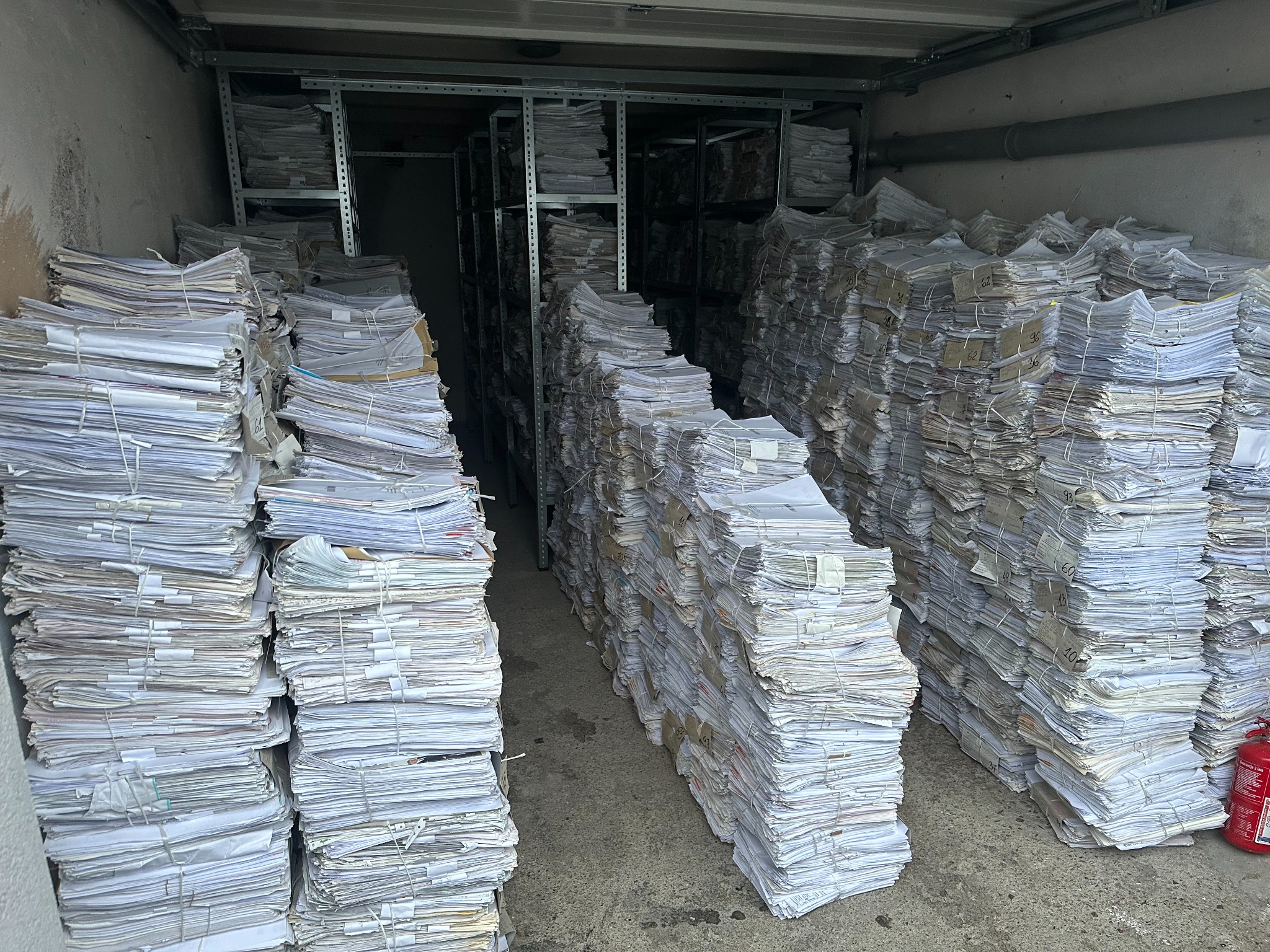 Stosy spiętych dokumentów ułożone w magazynie na regałach i podłodze, widoczna gaśnica w prawym dolnym rogu.