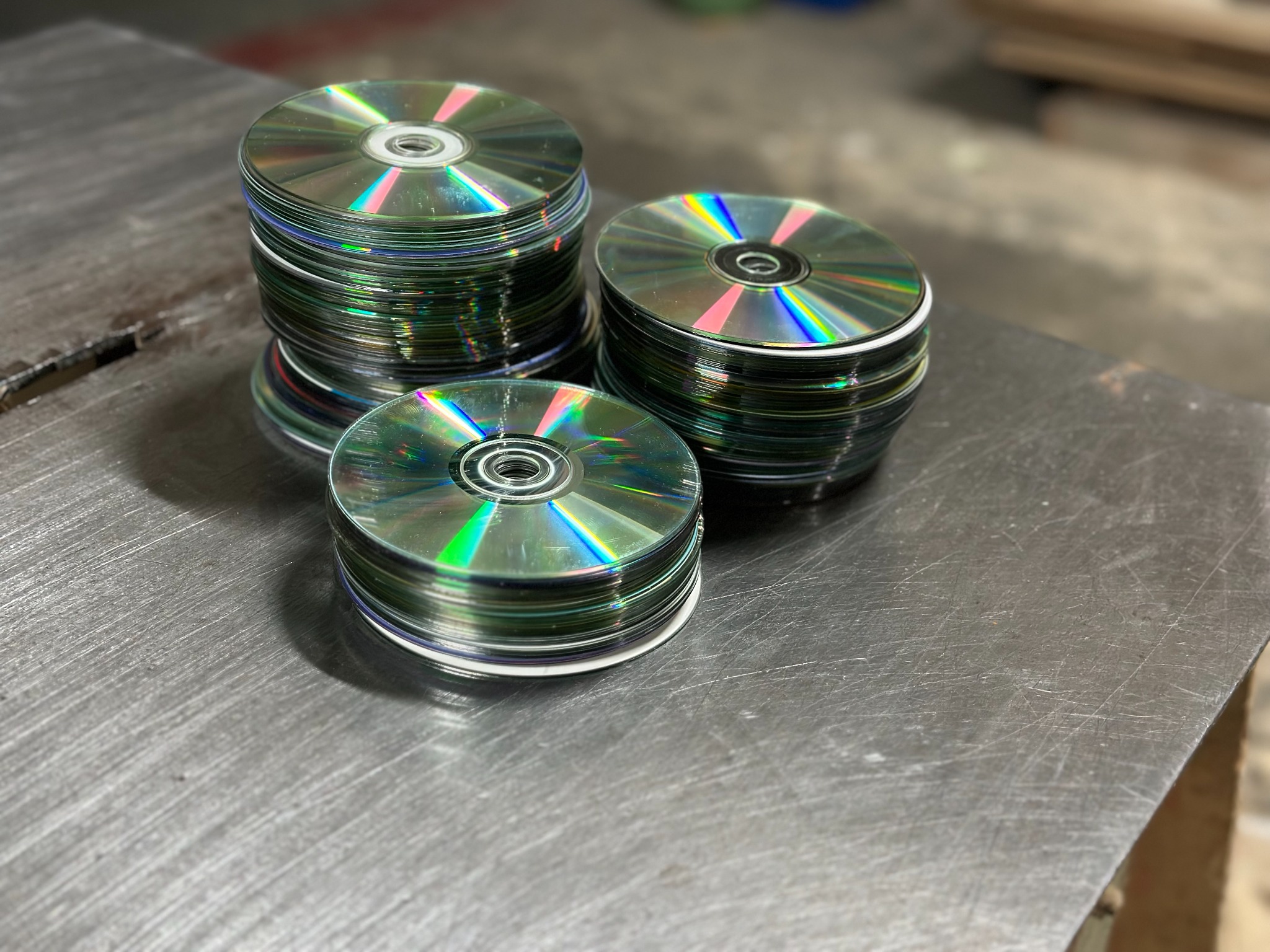 Trzy stosy srebrnych płyt CD na metalowej powierzchni. Widoczne tęczowe refleksy światła na powierzchni płyt.
