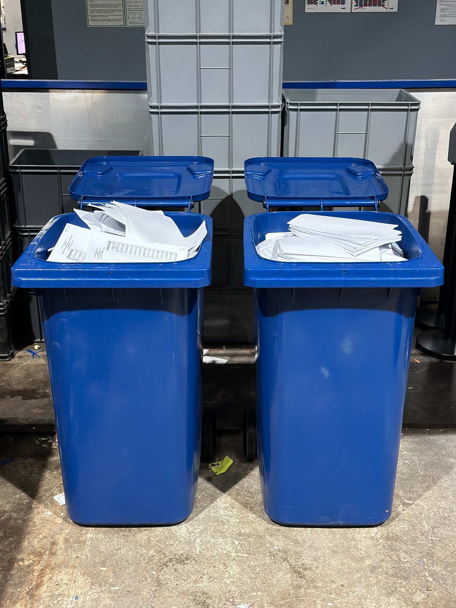 Dwa niebieskie pojemniki na kółkach wypełnione białymi dokumentami, z szarymi pojemnikami w tle, sugerujące archiwizację lub niszczenie dokumentów.