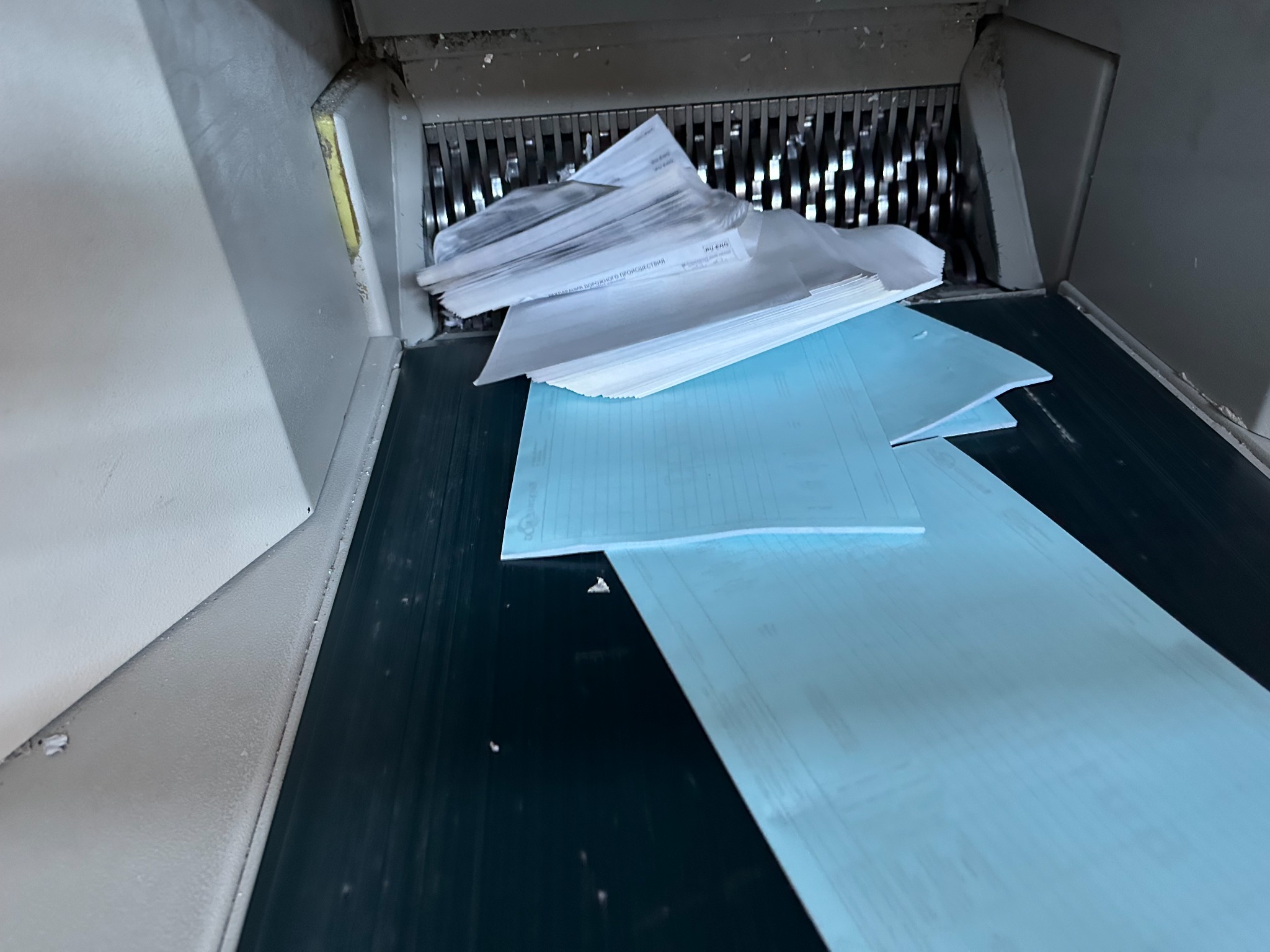 Stos białych i niebieskich dokumentów w podajniku niszczarki przemysłowej, widoczne ostrza tnące.