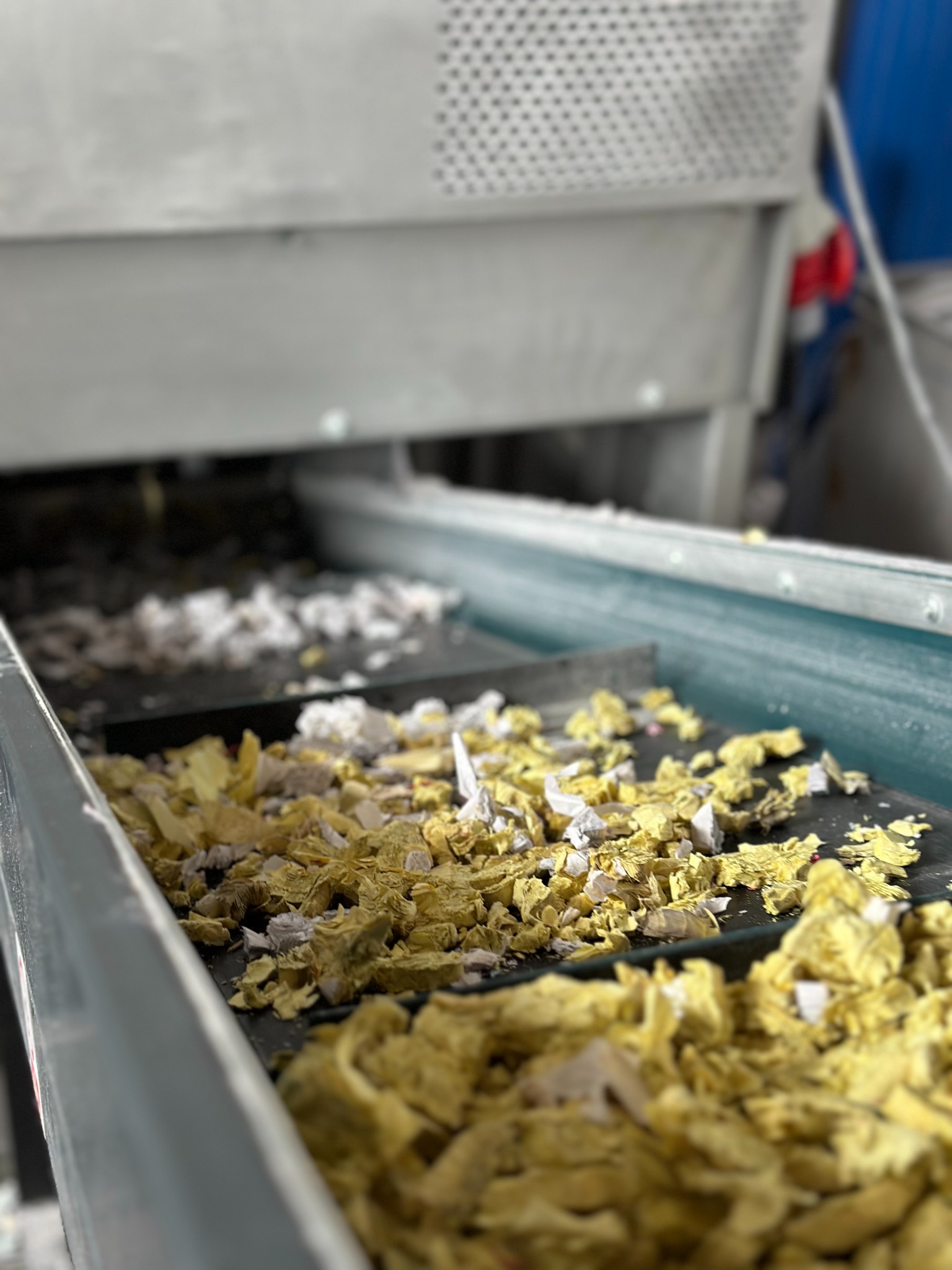 Fragment zniszczonych, pociętych dokumentów na taśmociągu wewnątrz maszyny do utylizacji papieru, widoczne resztki białych i żółtych kartek.