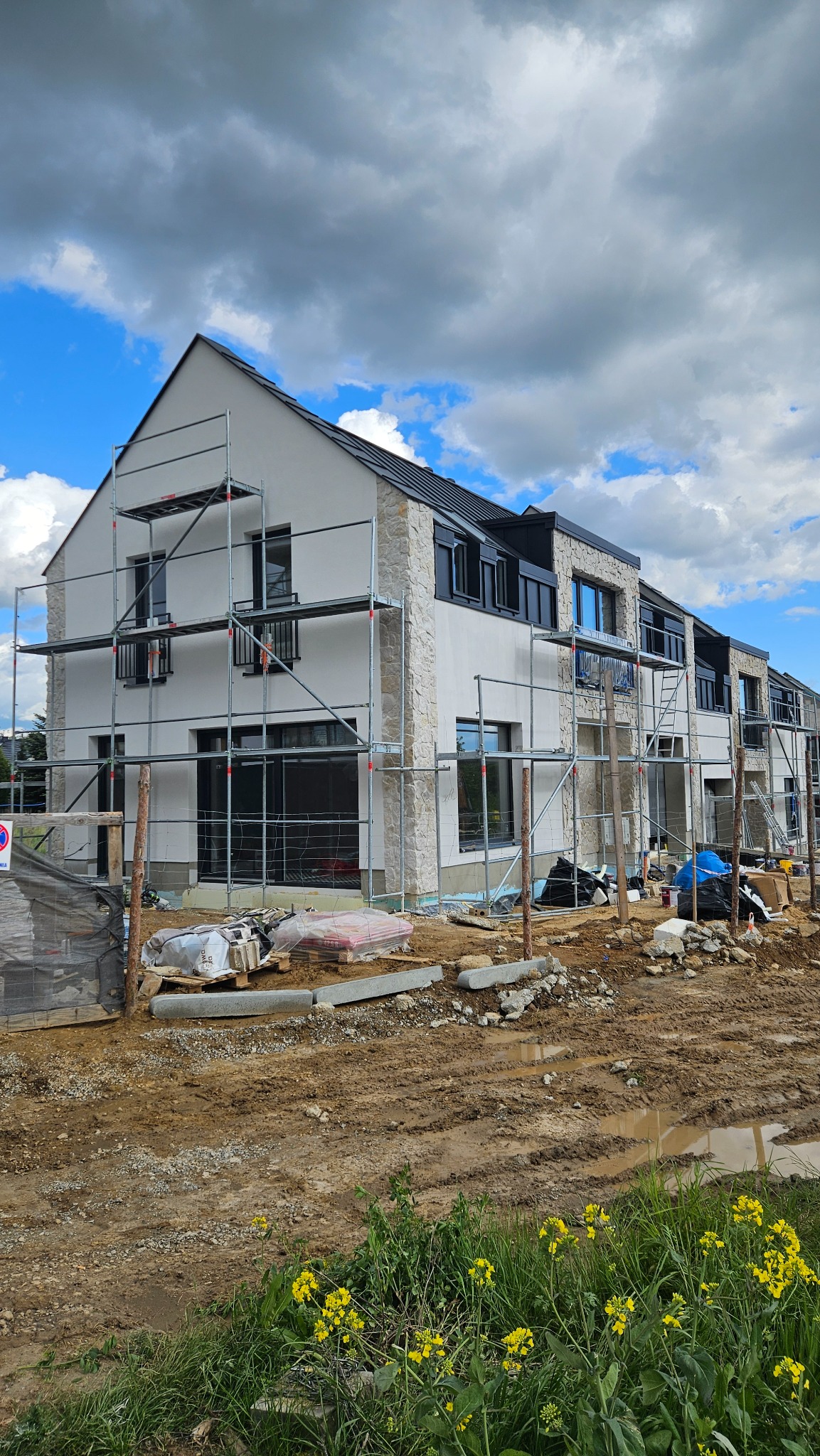 Nowoczesny budynek wielorodzinny w trakcie wykończenia elewacji, z widocznymi rusztowaniami i fragmentami kamiennej okładziny. Błękitne niebo z chmurami w tle.