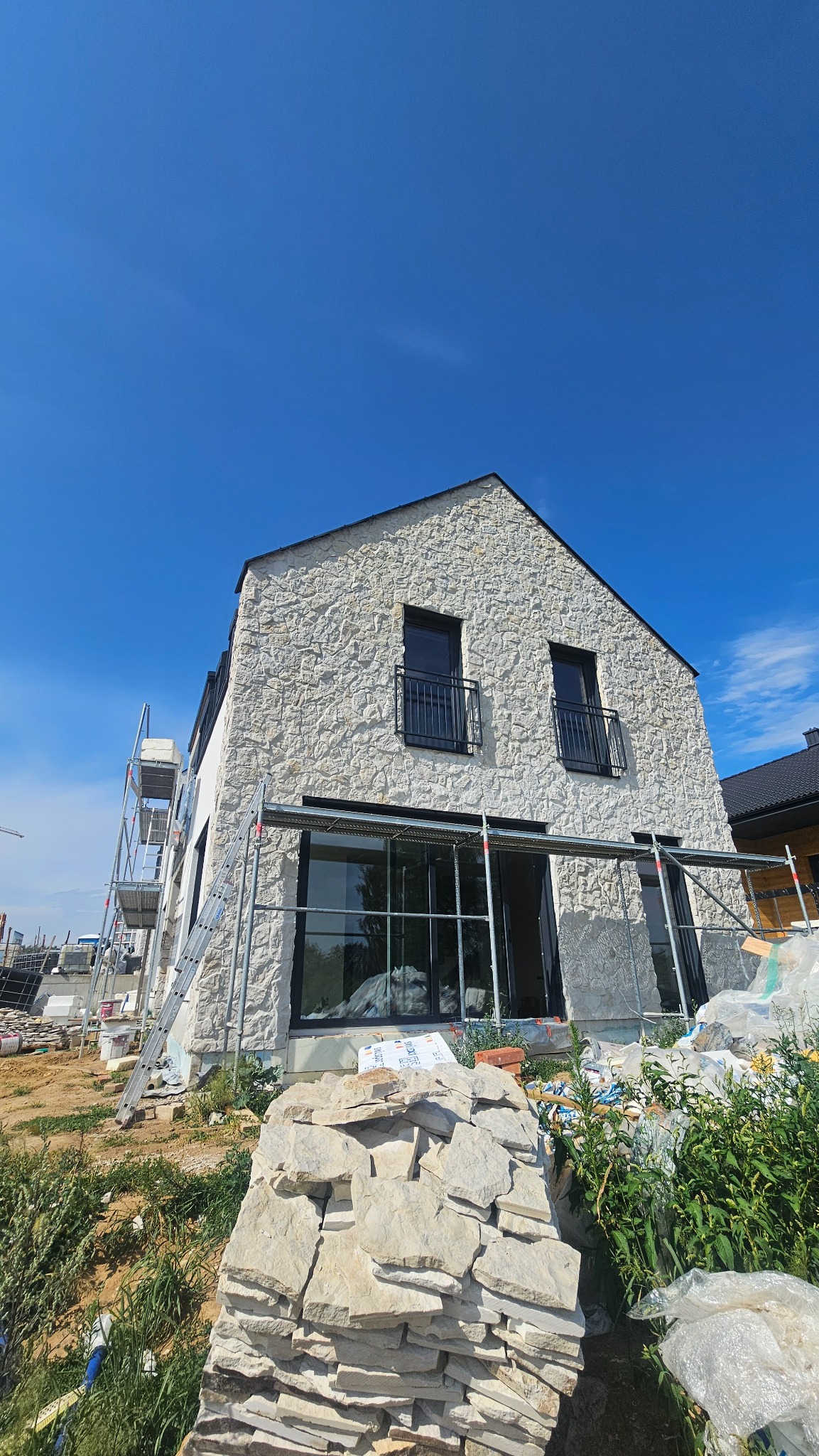 Dom z elewacją z kamienia naturalnego w trakcie wykańczania, widoczne rusztowania i stos kamieni. Błękitne niebo w tle.