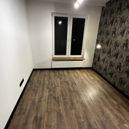Usługi remontowe Ewa Olszyńska - Pusty pokój po remoncie z panelami podłogowymi w odcieniach brązu, białe ściany z czarnymi listwami przypodłogowymi, okno z drewnianym parapetem i grzejnikiem, jedna ściana z tapetą imitującą...