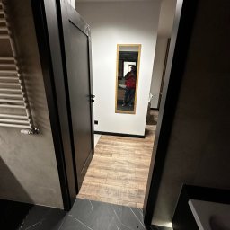 Usługi remontowe Ewa Olszyńska - Częściowo otwarte czarne drzwi do łazienki z czarną marmurową podłogą z białymi żyłami i drewnopodobną podłogą w głębi, widoczne lustro z odbiciem osoby.