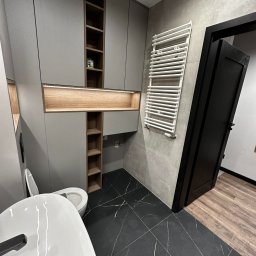 Usługi remontowe Ewa Olszyńska - Nowoczesna łazienka z szarą zabudową meblową, podświetlanymi półkami, czarną marmurową podłogą i białym grzejnikiem drabinkowym, widok na otwarte czarne drzwi.