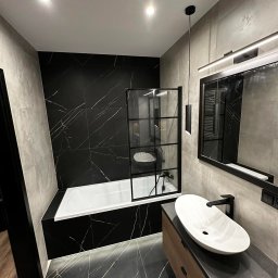 Usługi remontowe Ewa Olszyńska - Nowoczesna łazienka z czarnymi płytkami imitującymi marmur, białą wanną za czarną, szklaną ścianką, owalną umywalką nablatową i dużym lustrem z oświetleniem LED.