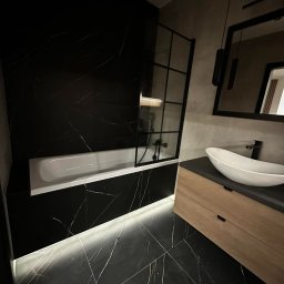 Usługi remontowe Ewa Olszyńska - Nowoczesna łazienka w czarno-białej kolorystyce z akcentami drewna: czarne marmurowe płytki z białymi żyłami, biała wanna z parawanem, umywalka nablatowa na drewnianej szafce, oświetlenie LED...