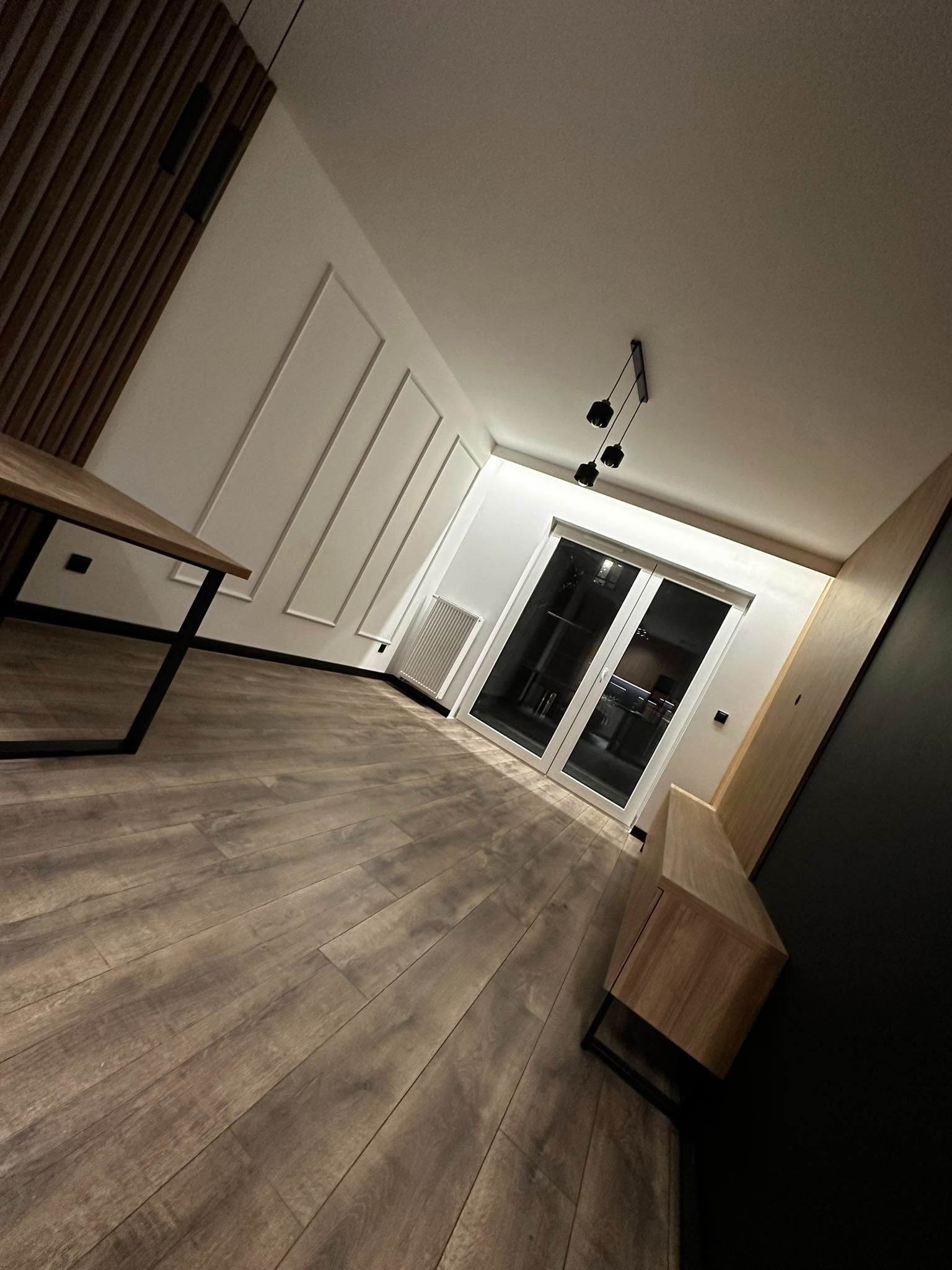Nowoczesne wnętrze z drewnianą podłogą, minimalistycznymi listwami ściennymi, drewnianą zabudową i stolikiem na metalowych nogach, oświetlone trzema czarnymi lampami wiszącymi.