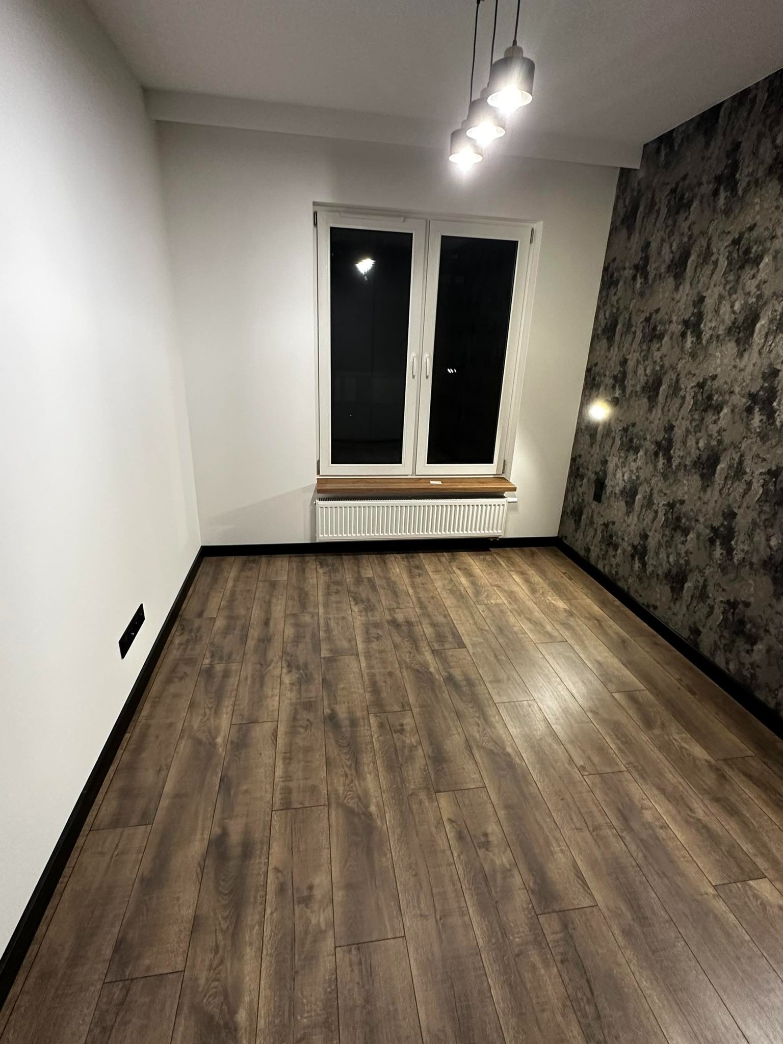 Pusty pokój po remoncie z panelami podłogowymi w odcieniach brązu, białe ściany z czarnymi listwami przypodłogowymi, okno z drewnianym parapetem i grzejnikiem, jedna ściana z tapetą imitującą...