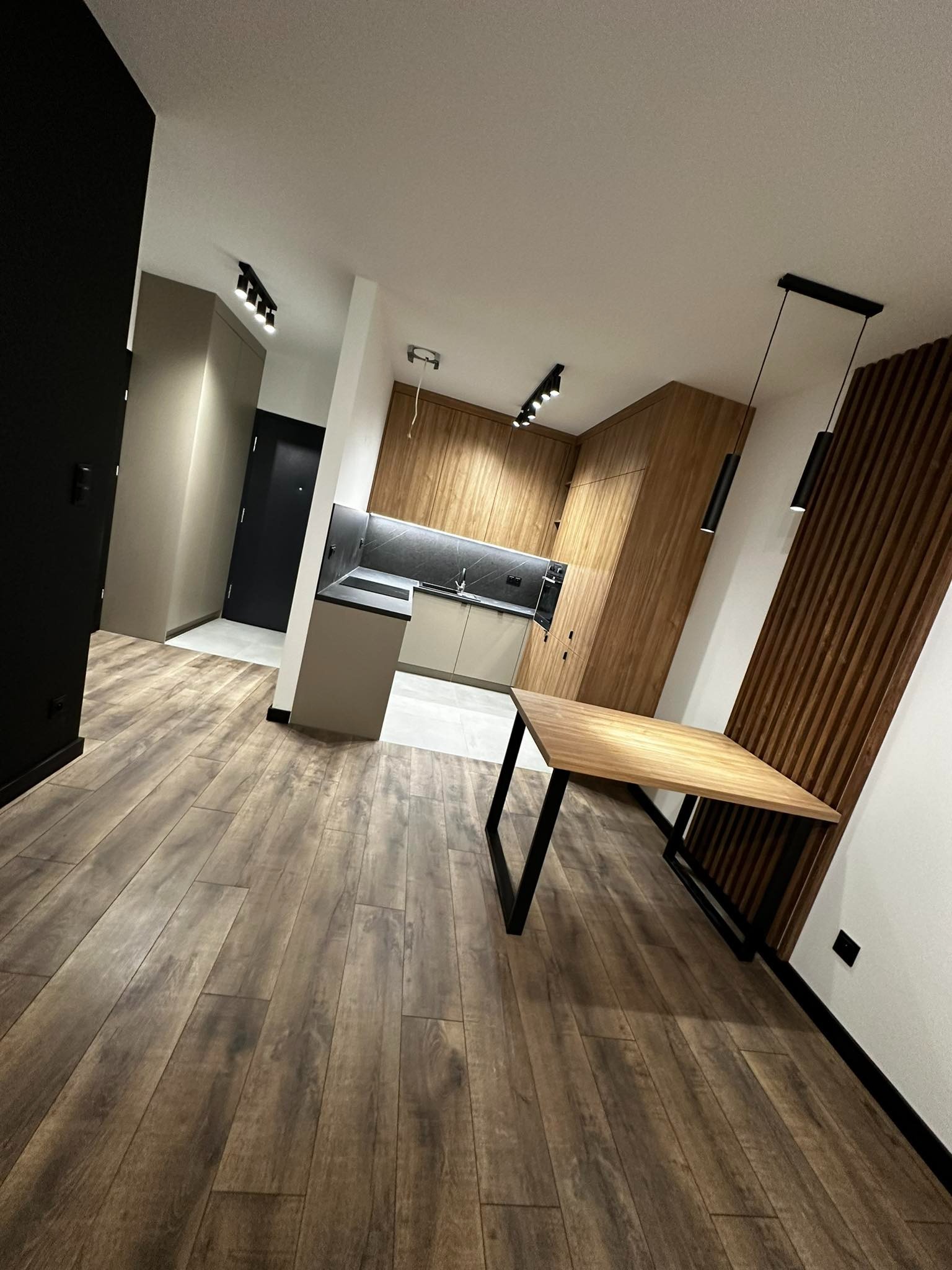 Nowoczesne wnętrze: drewniana podłoga, zabudowa kuchenna z drewnianymi frontami i czarnym blatem, stół z drewnianym blatem i czarnymi nogami, dekoracyjna ściana z drewnianych listew, oświetlenie...