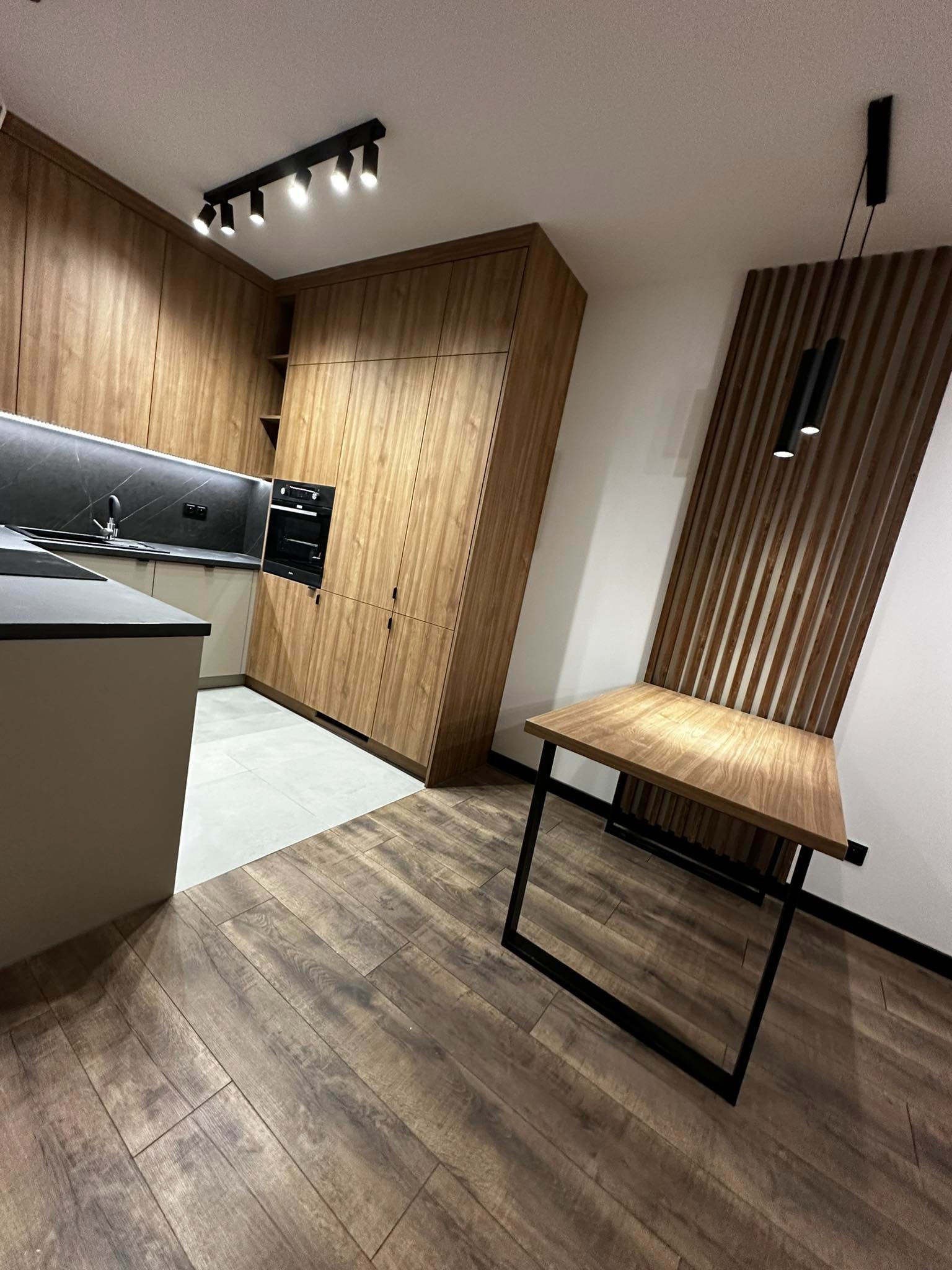 Nowoczesna kuchnia z drewnianymi szafkami i blatem w Warszawie, uzupełniona o drewniany panel dekoracyjny i stół z czarnymi nogami.