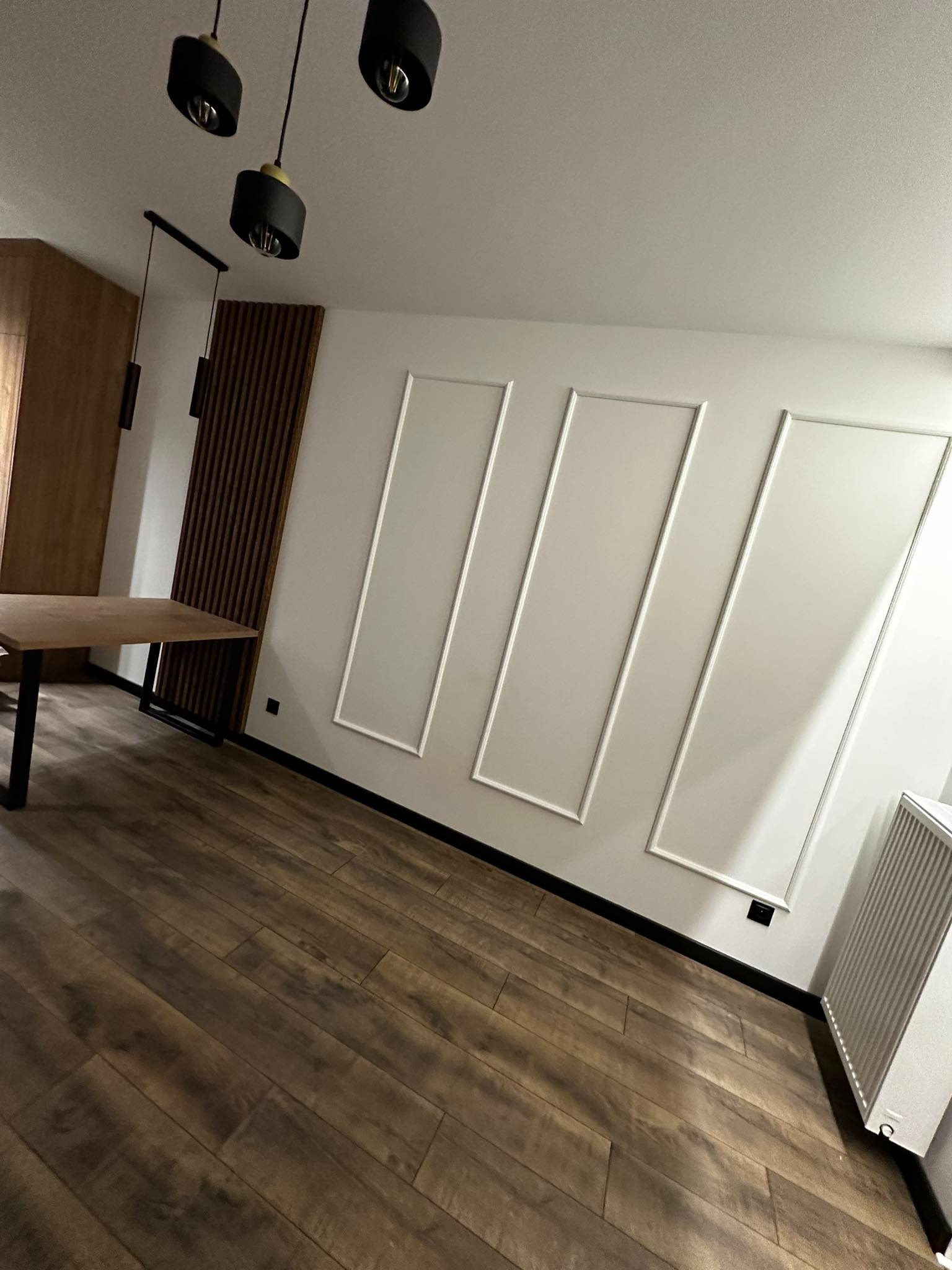 Nowoczesne wnętrze z drewnianą podłogą, białymi ścianami z dekoracyjnymi listwami, drewnianą ścianką działową i wiszącymi lampami.