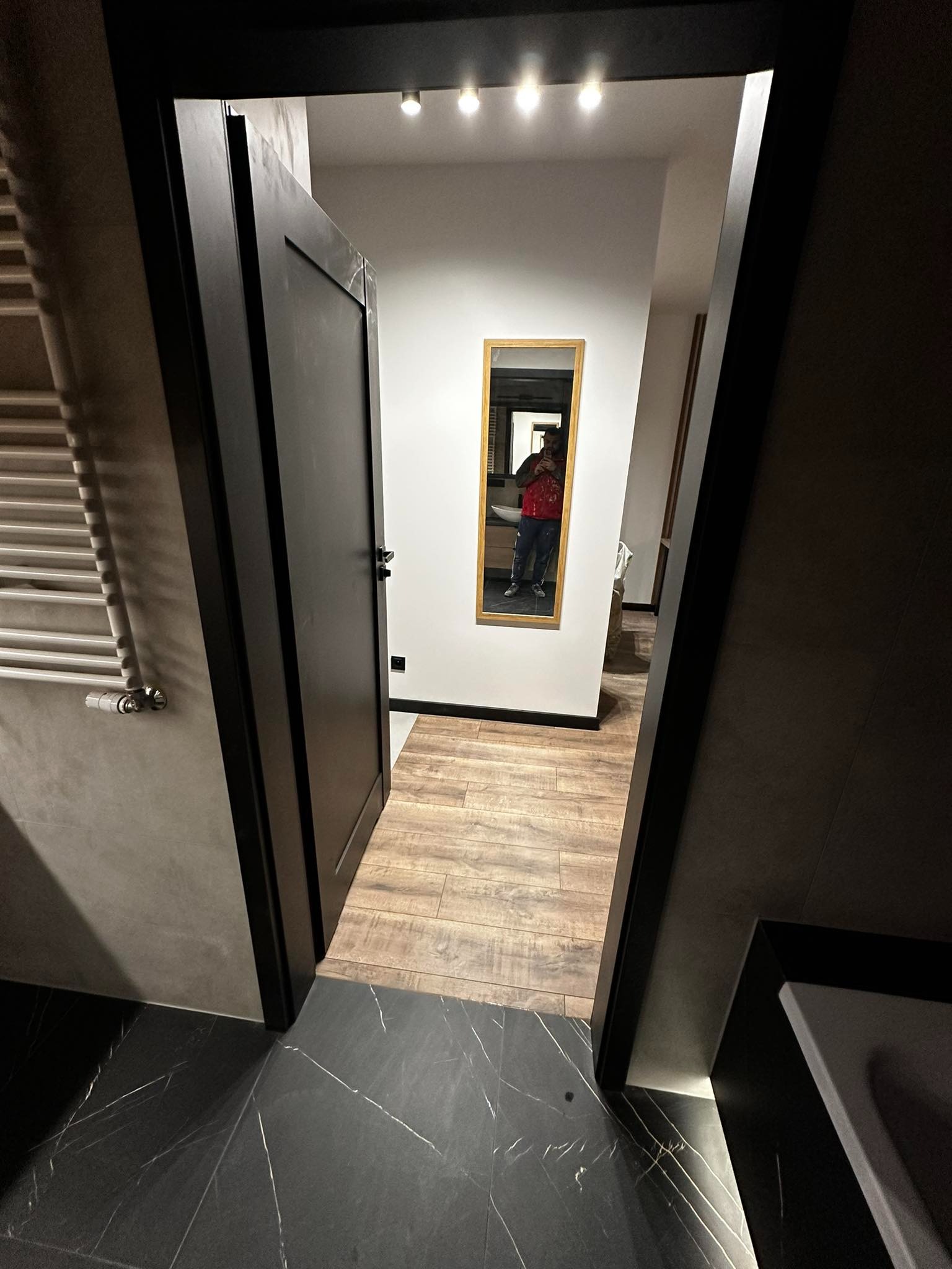 Częściowo otwarte czarne drzwi do łazienki z czarną marmurową podłogą z białymi żyłami i drewnopodobną podłogą w głębi, widoczne lustro z odbiciem osoby.