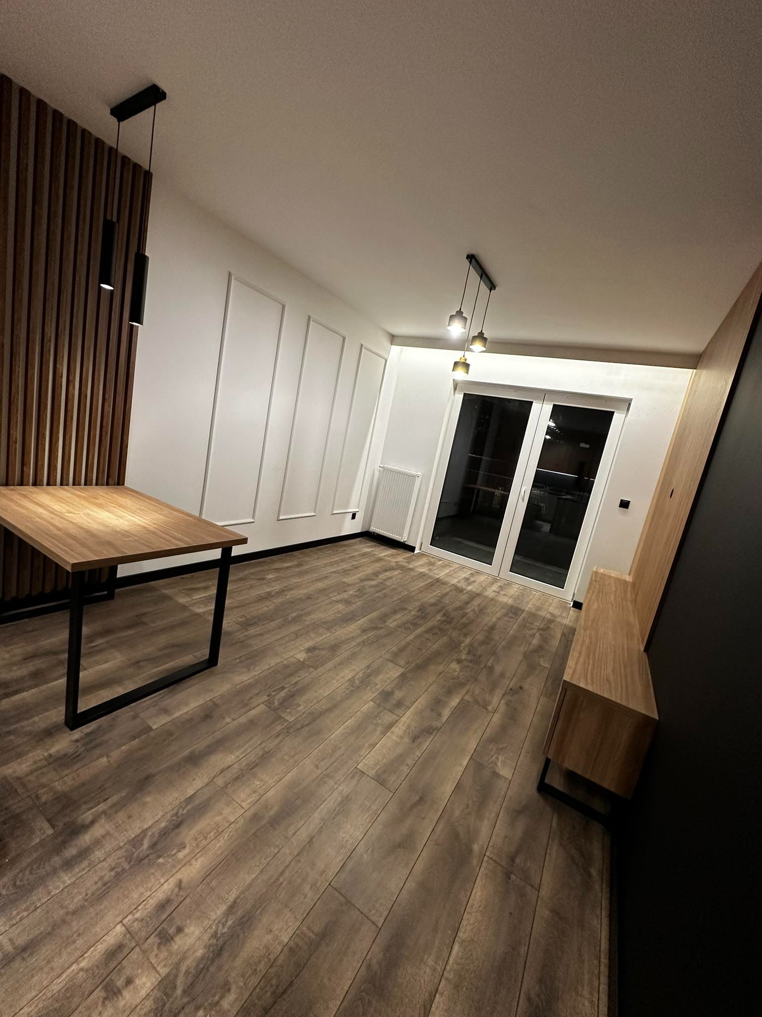 Nowoczesne wnętrze z drewnianą podłogą, panele ścienne, stół z metalowymi nogami, oświetlenie punktowe i przesuwne drzwi balkonowe.