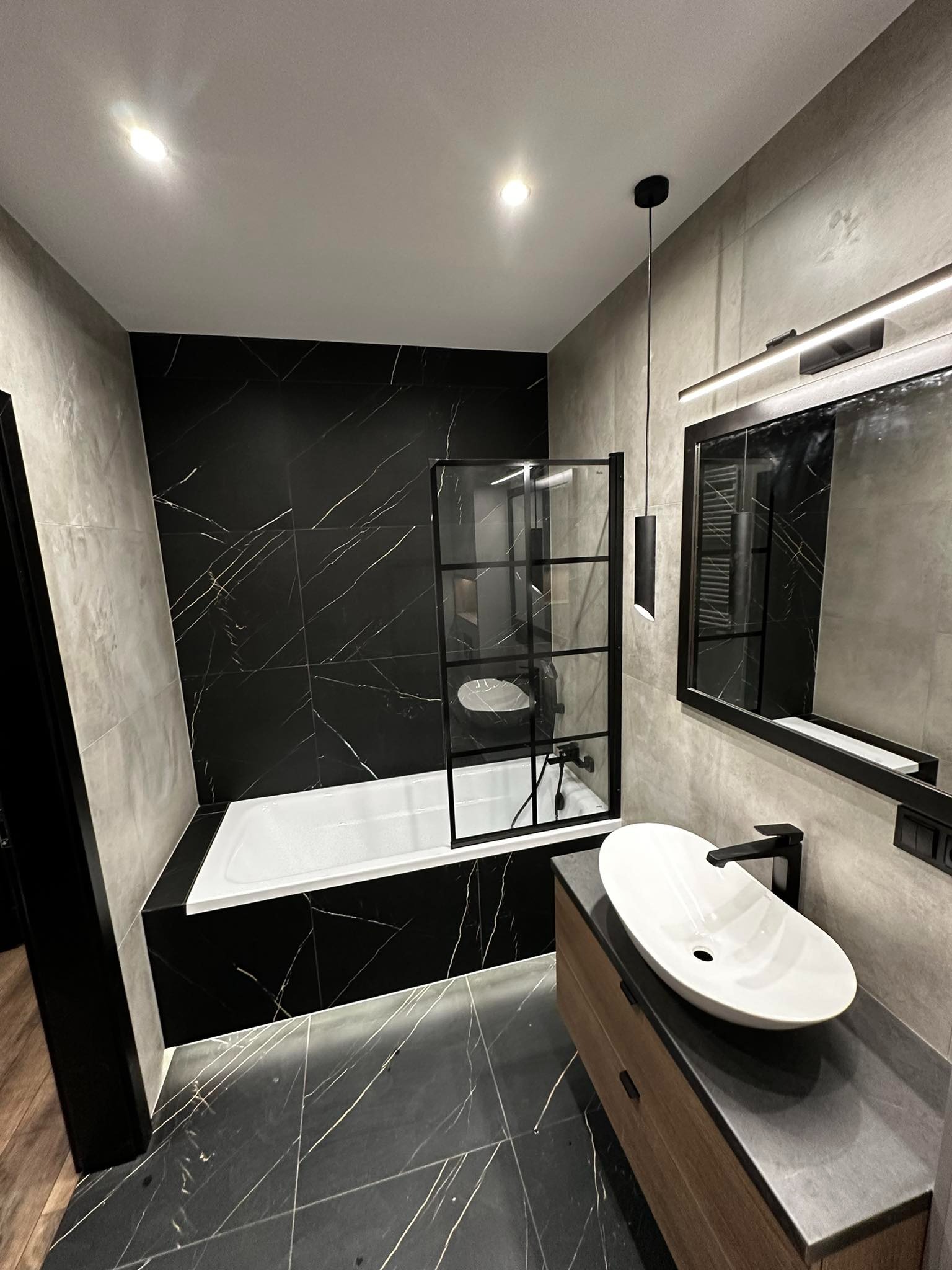 Nowoczesna łazienka z czarnymi płytkami imitującymi marmur, białą wanną z czarną szybą prysznicową, owalną umywalką nablatową i drewnianą szafką pod umywalkę, oświetlona lampą wiszącą i listwą LED...