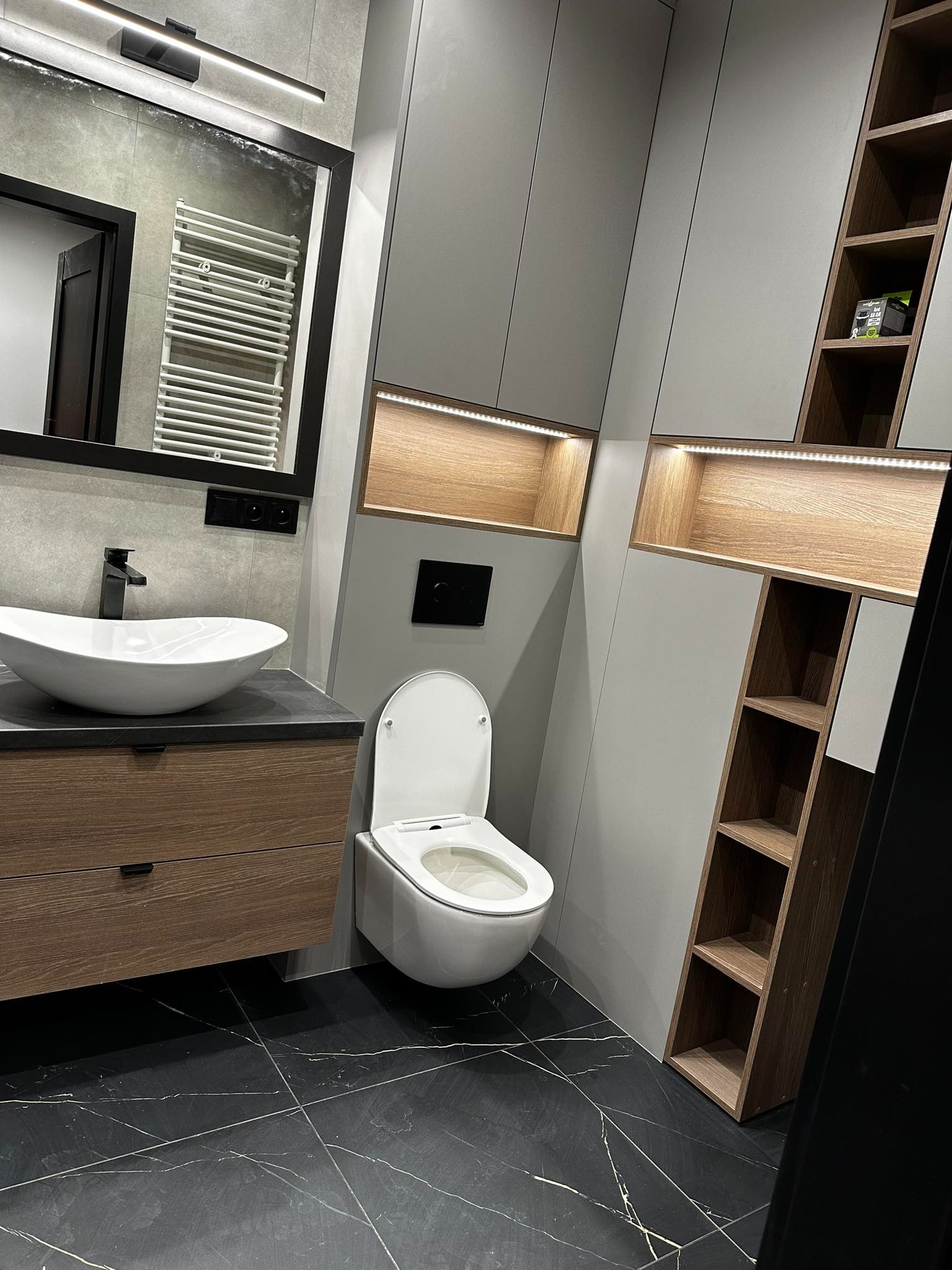 Nowoczesna łazienka z umywalką nablatową, toaletą podwieszaną i szafkami w odcieniach szarości i drewna, czarna marmurowa podłoga.