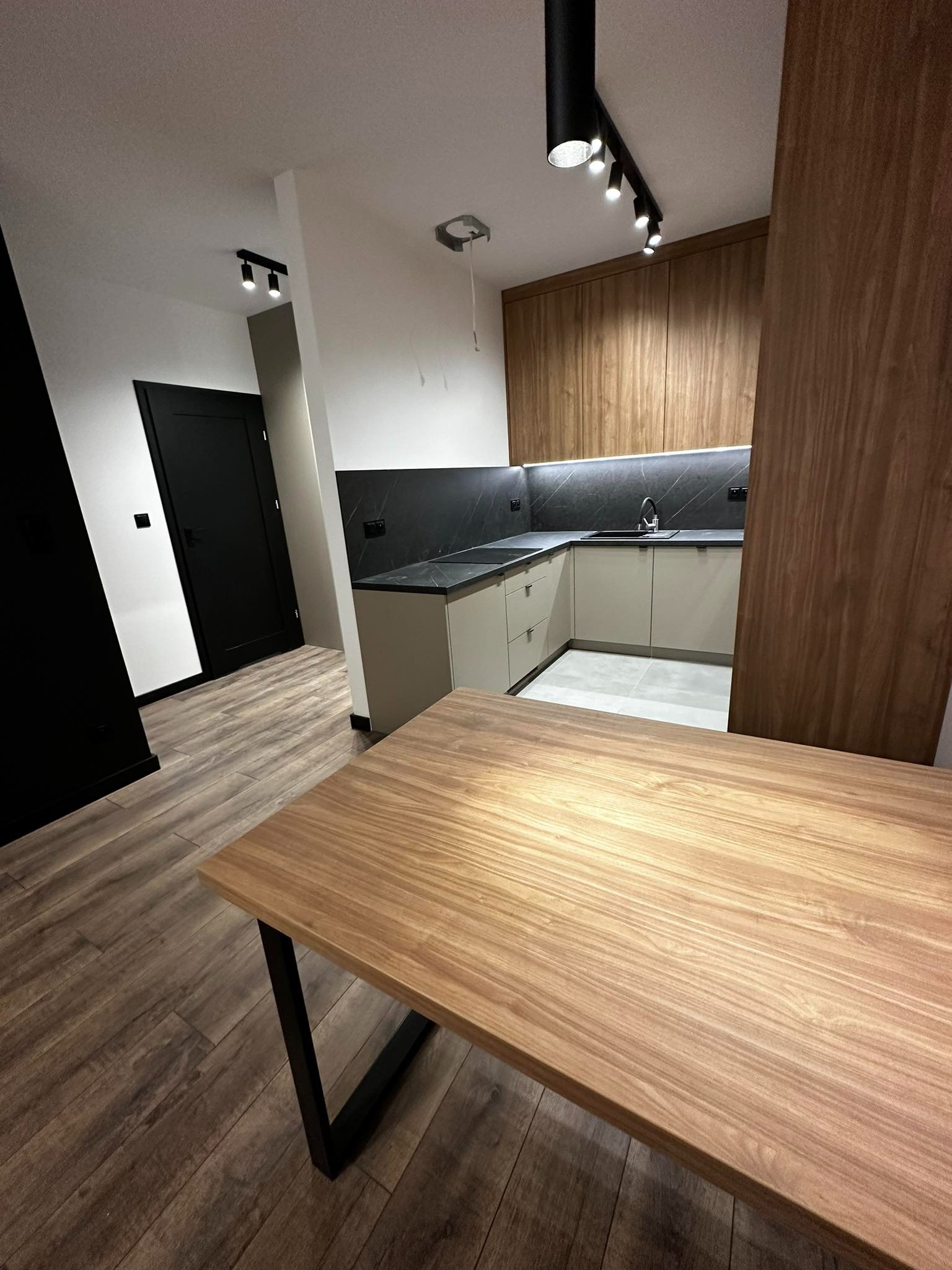 Nowoczesna kuchnia z drewnianymi szafkami górnymi, szarym blatem i oświetleniem punktowym, widziana z perspektywy blatu stołu z drewnianym blatem i metalowymi nogami.