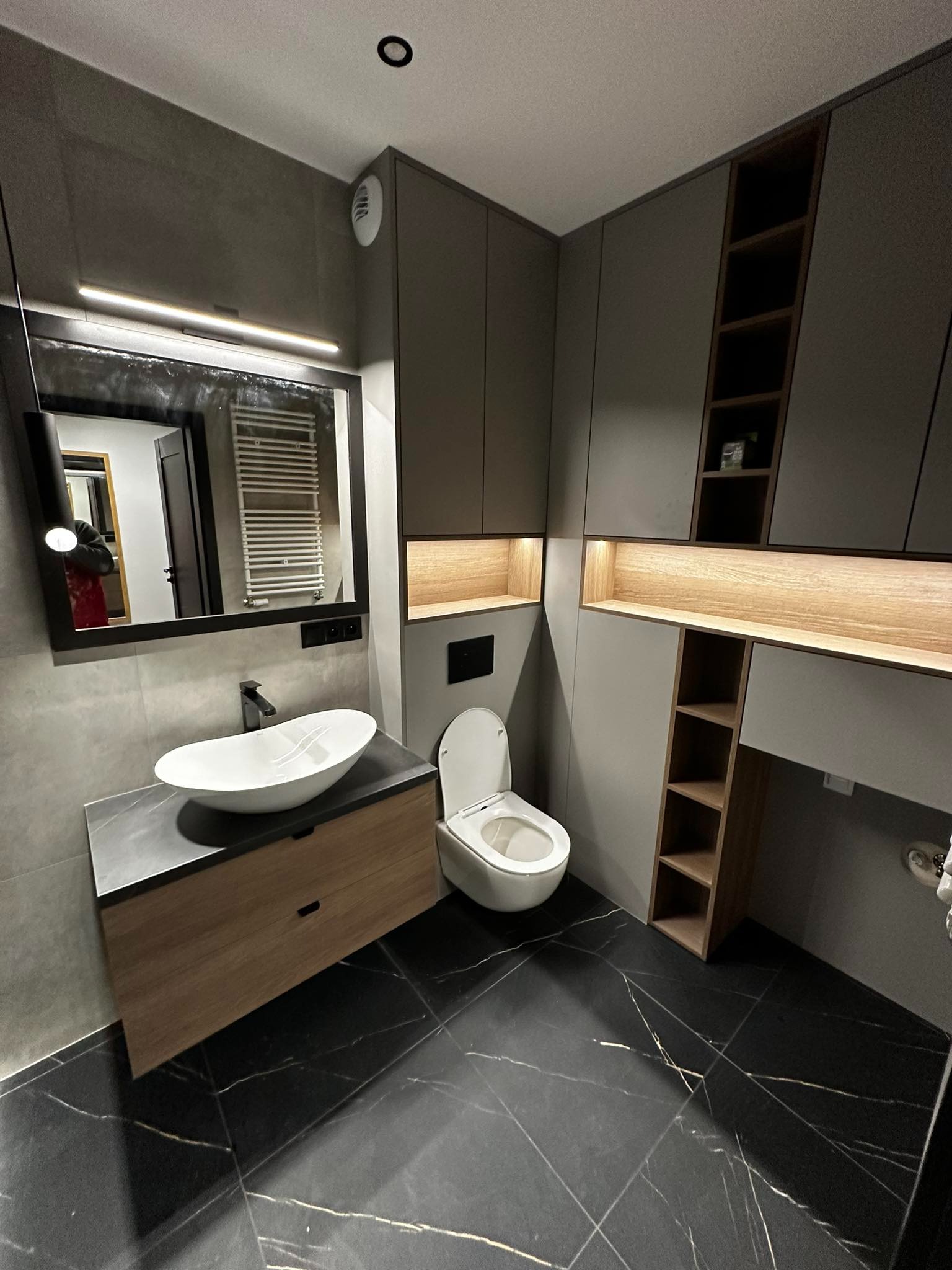 Nowoczesna łazienka z czarną marmurową podłogą, owalną umywalką na drewnianej szafce, szarą zabudową meblową z oświetleniem LED i dużym lustrem z czarną ramą.