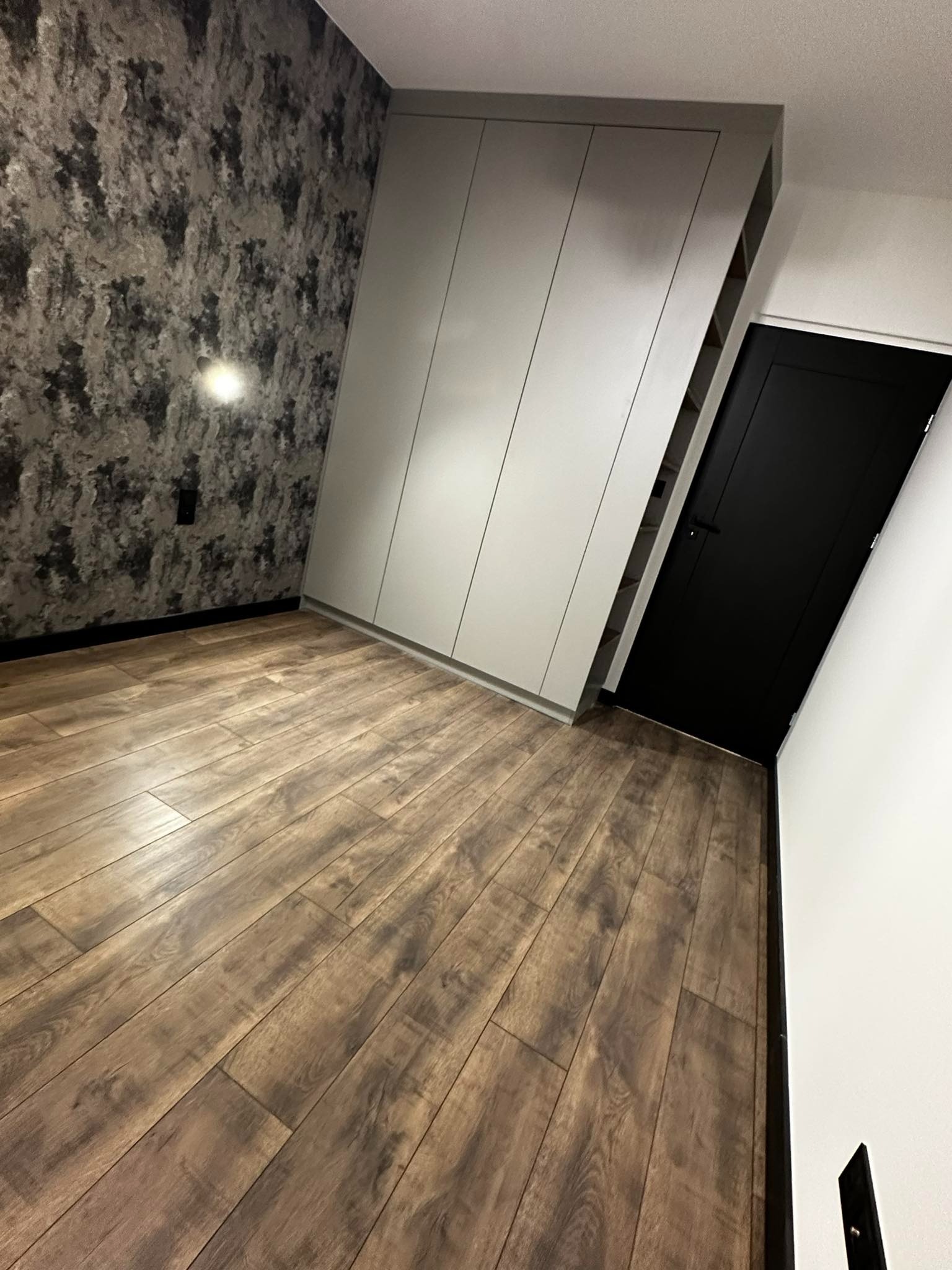 Pokój po odnowieniu: drewnopodobna podłoga, ściana z tapetą imitującą kamień, szara szafa wnękowa i czarne drzwi.
